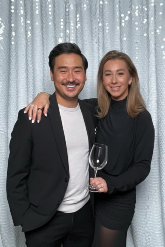 A man and woman standing close together with their arms around each other in front of a light-colored curtain. The man is wearing a black blazer and white shirt, and the woman is wearing a black dress and holding a wine glass.