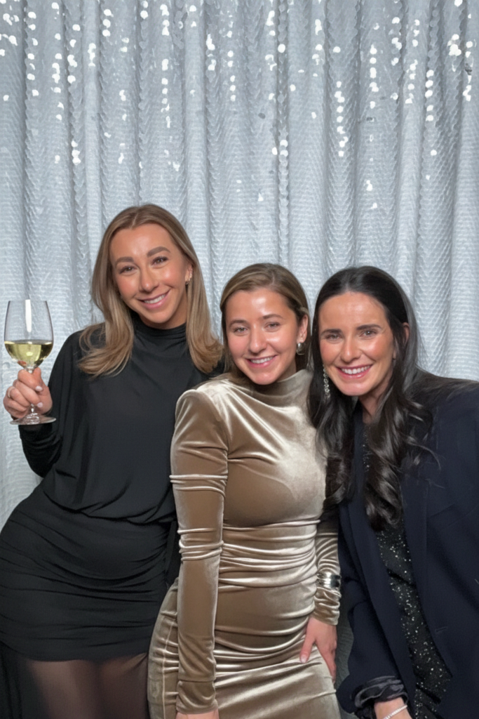Three women smiling and posing together indoors in front of a light-colored curtain. The woman on the left is holding a glass of white wine, the woman in the middle is wearing a shiny, gold dress, and the woman on the right has dark hair and is dress