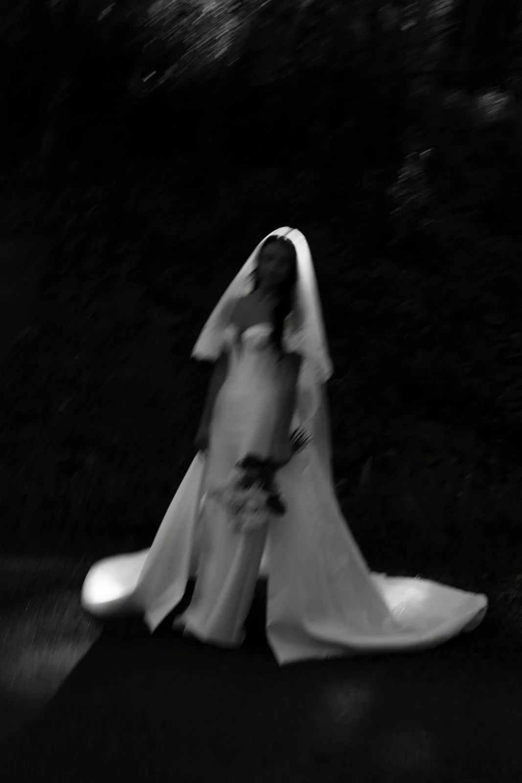 A black and white photo of a woman dressed as a bride, standing outdoors with a dark background, holding a bouquet, with a blurry focus.