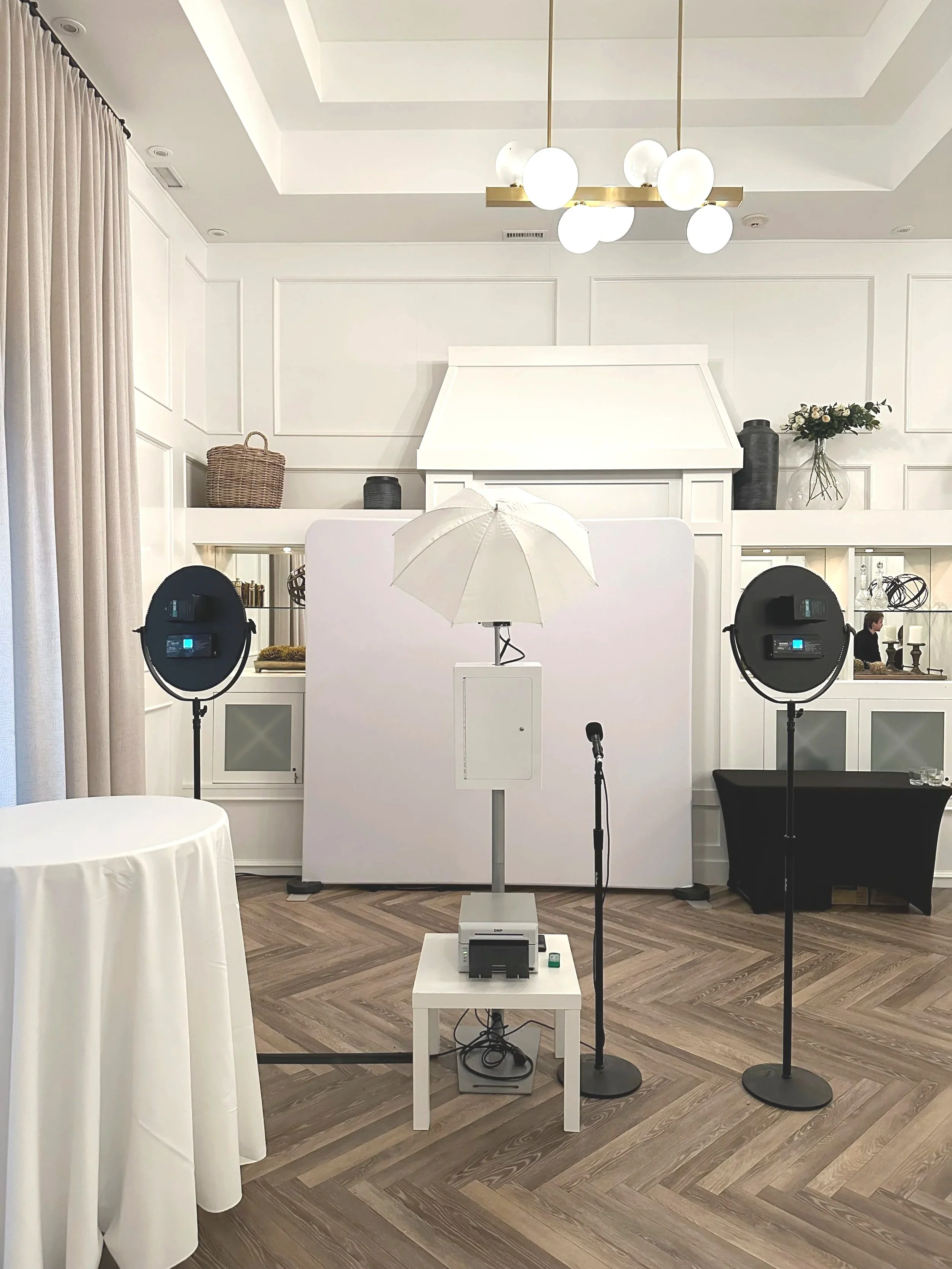 Photography setup with a white backdrop, two studio lights, a parasol, and a microphone in a well-lit indoor space with elegant decor and wooden flooring.