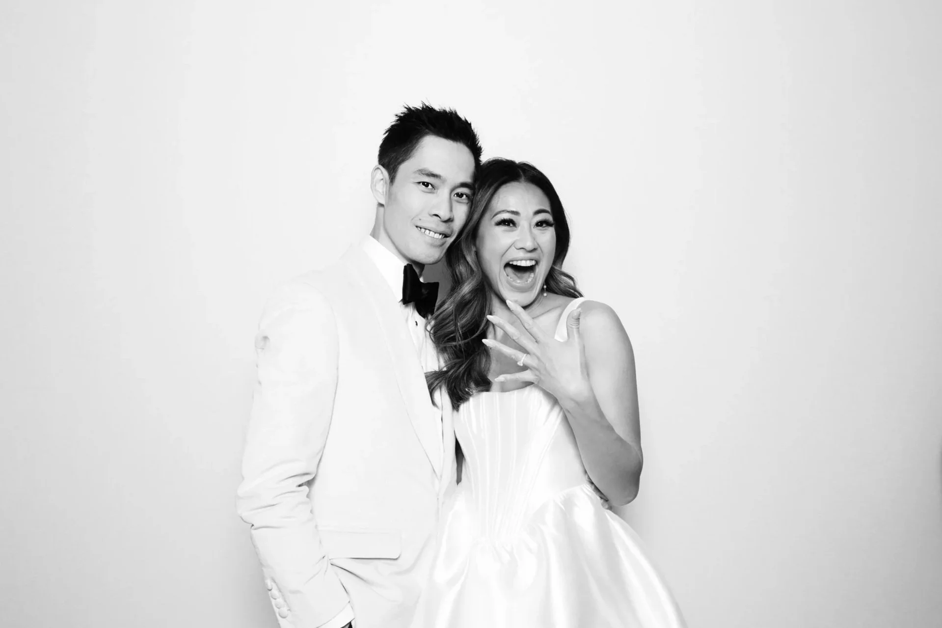 A black and white photograph of a happy couple at their wedding, with the woman wearing a wedding dress and showing off her engagement ring, and the man dressed in a white suit with a bow tie.