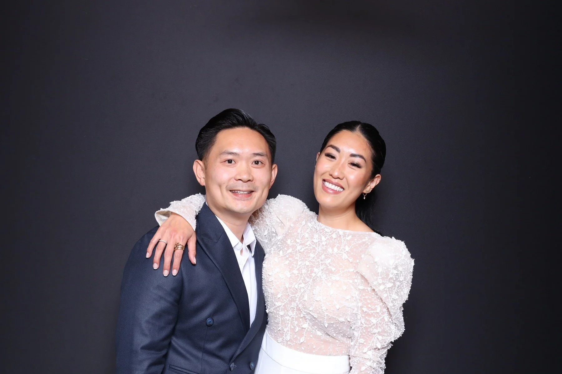 A happy couple smiling, with the woman wearing a white textured dress and the man in a dark suit, against a black background.