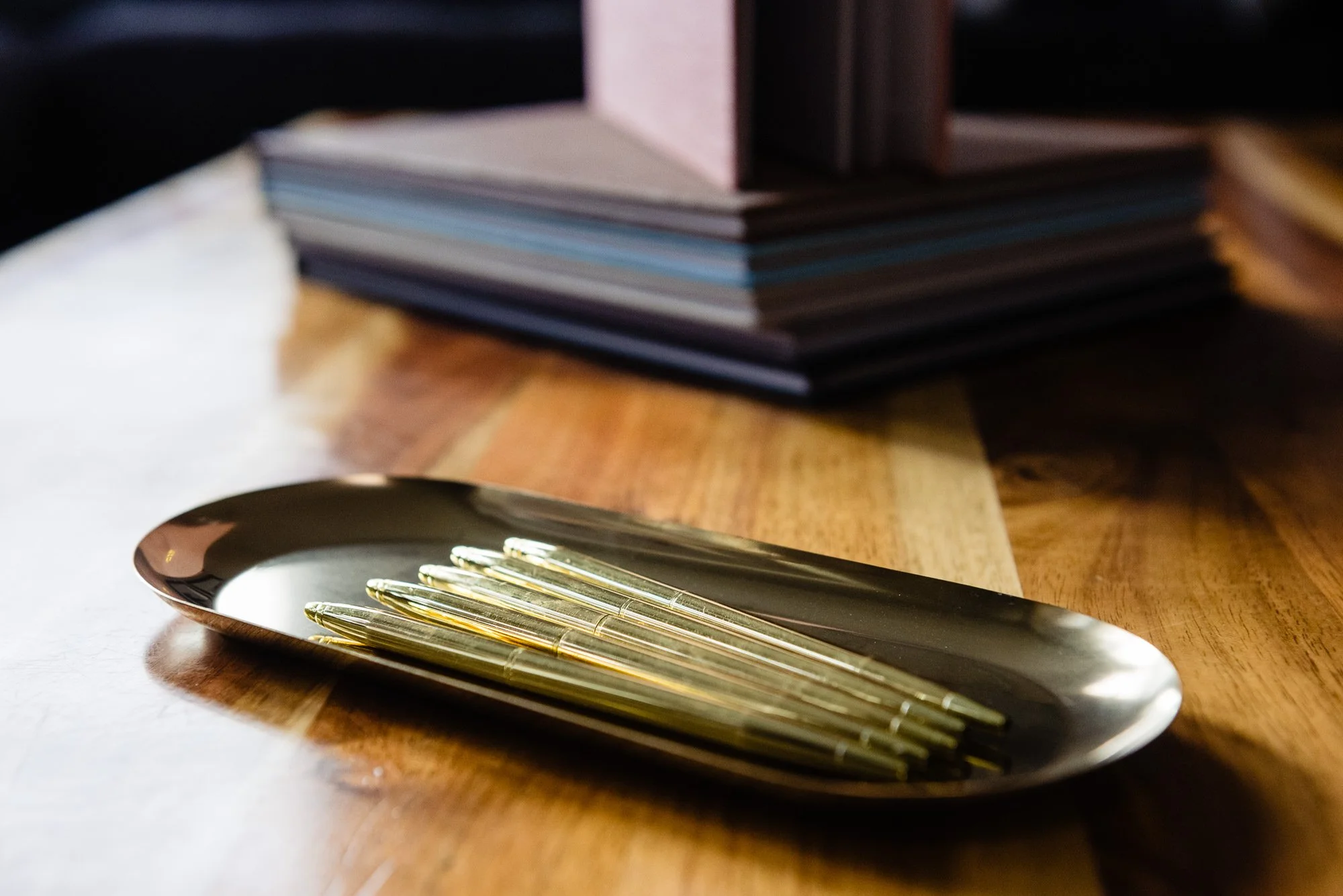 A black oval plate with gold pens on it, placed on a wooden table, with a blurred stack of papers or notebooks in the background.