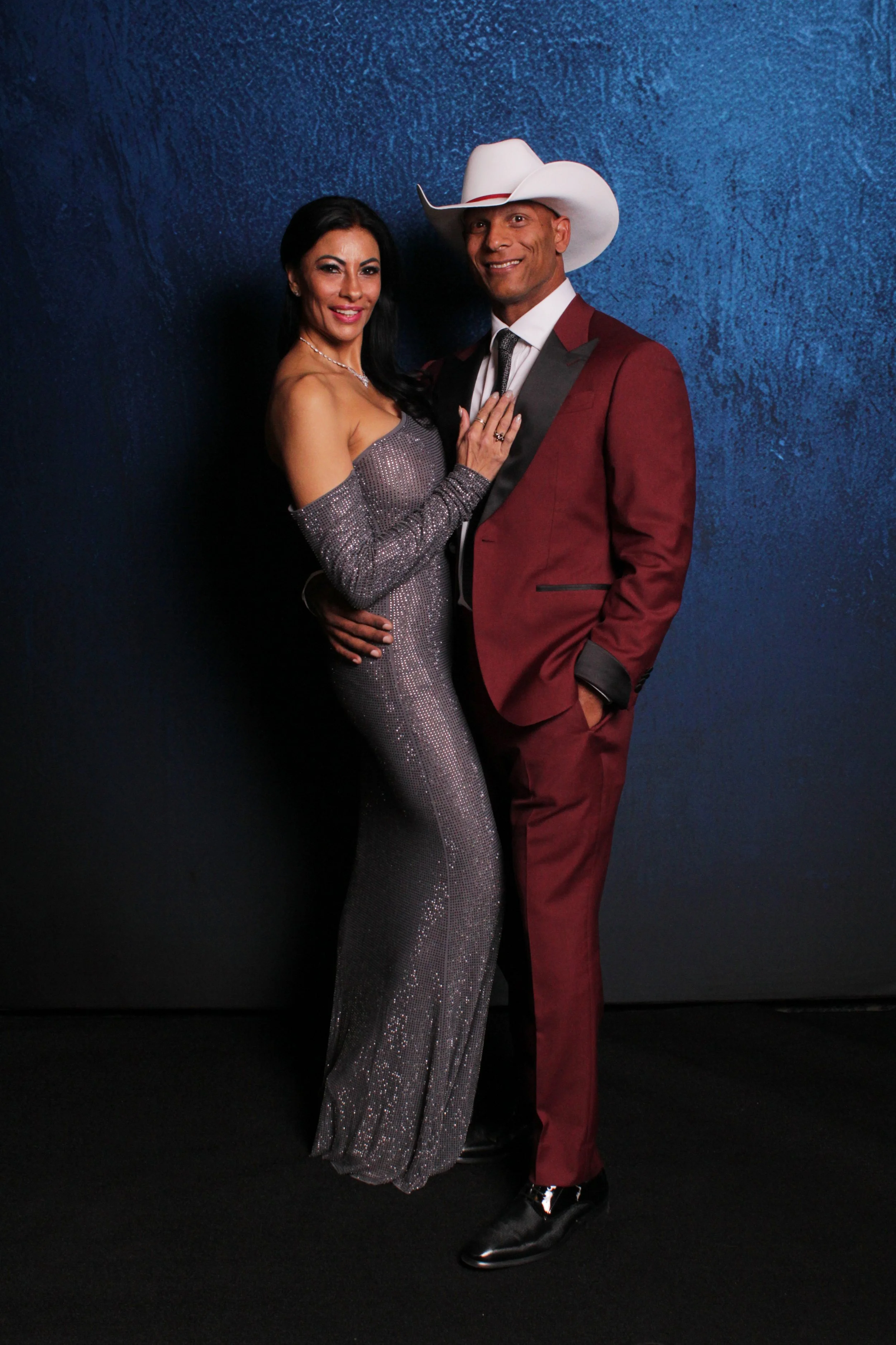 A woman in a silver sequined gown and a man in a burgundy suit with a white cowboy hat posing together against a dark blue textured background.