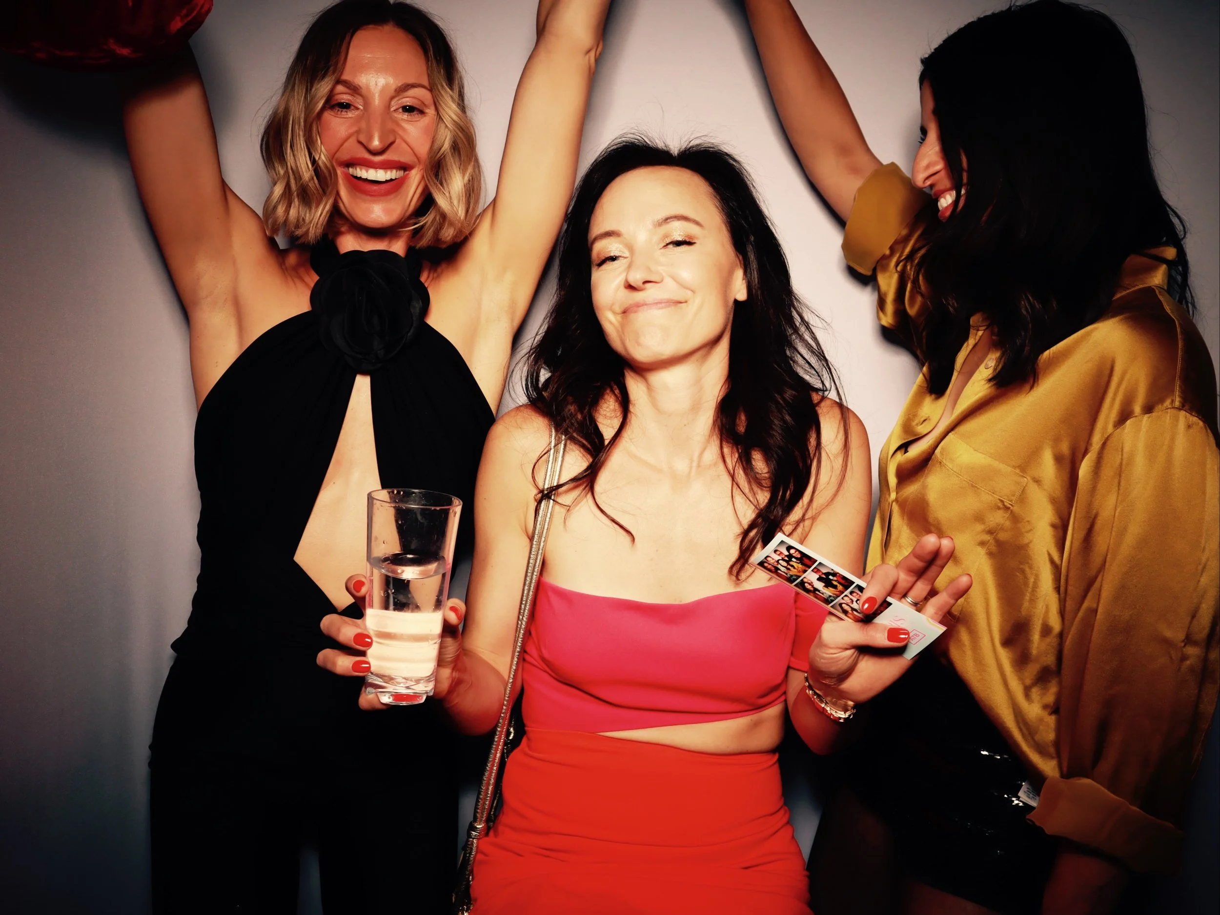 Three women celebrating, one holding a glass of water and photo booth strips, smiling and enjoying a party.