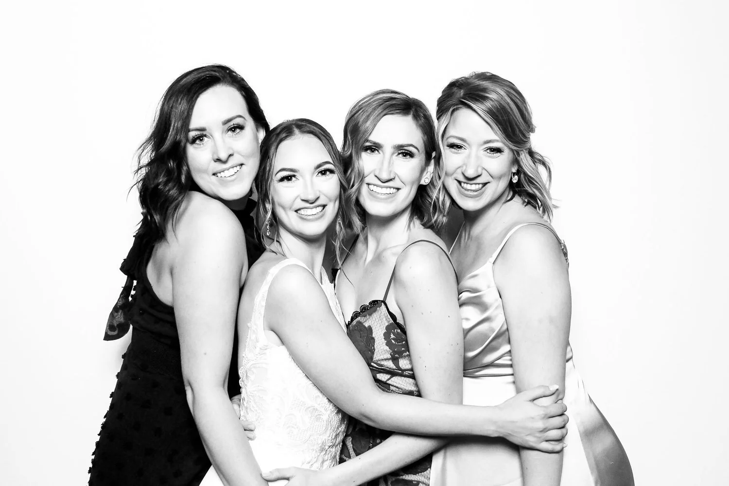 Four women smiling closely together in a black-and-white photo, dressed in elegant dresses.