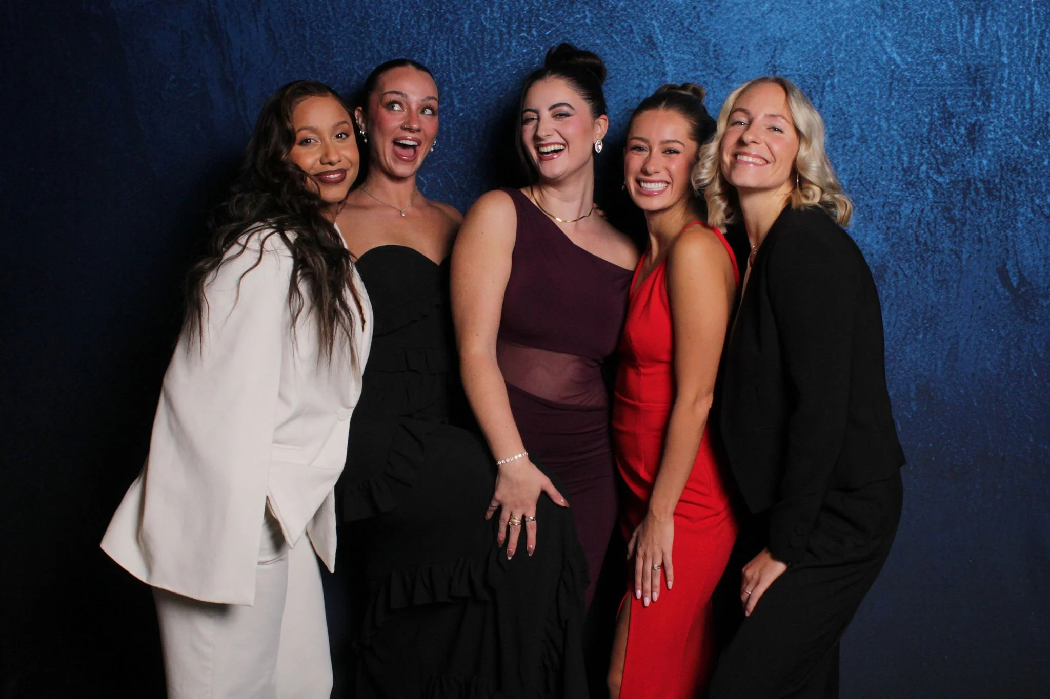 Five women dressed in formal attire standing close together and smiling against a textured blue background.