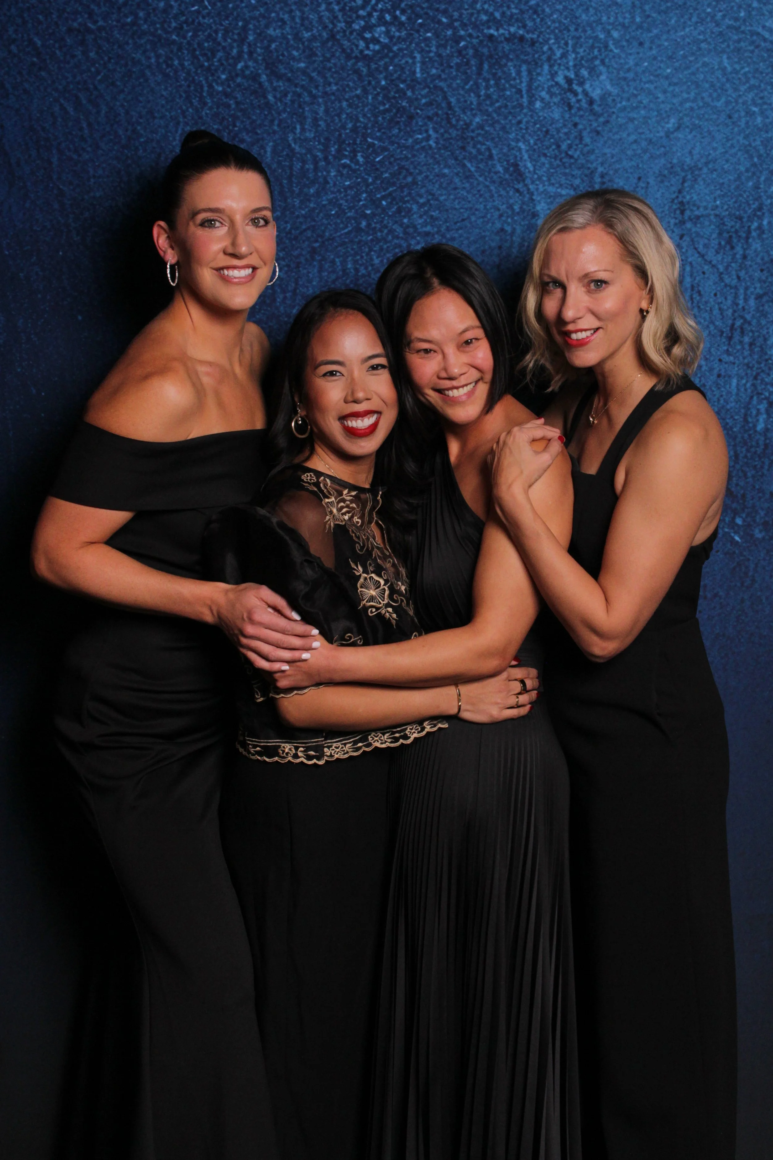 Four women in black dresses smiling and hugging against a blue textured backdrop.