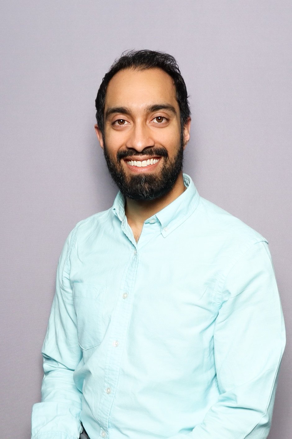 A man with dark hair, beard, and light skin wearing a light blue button-up shirt, smiling and standing against a gray background.
