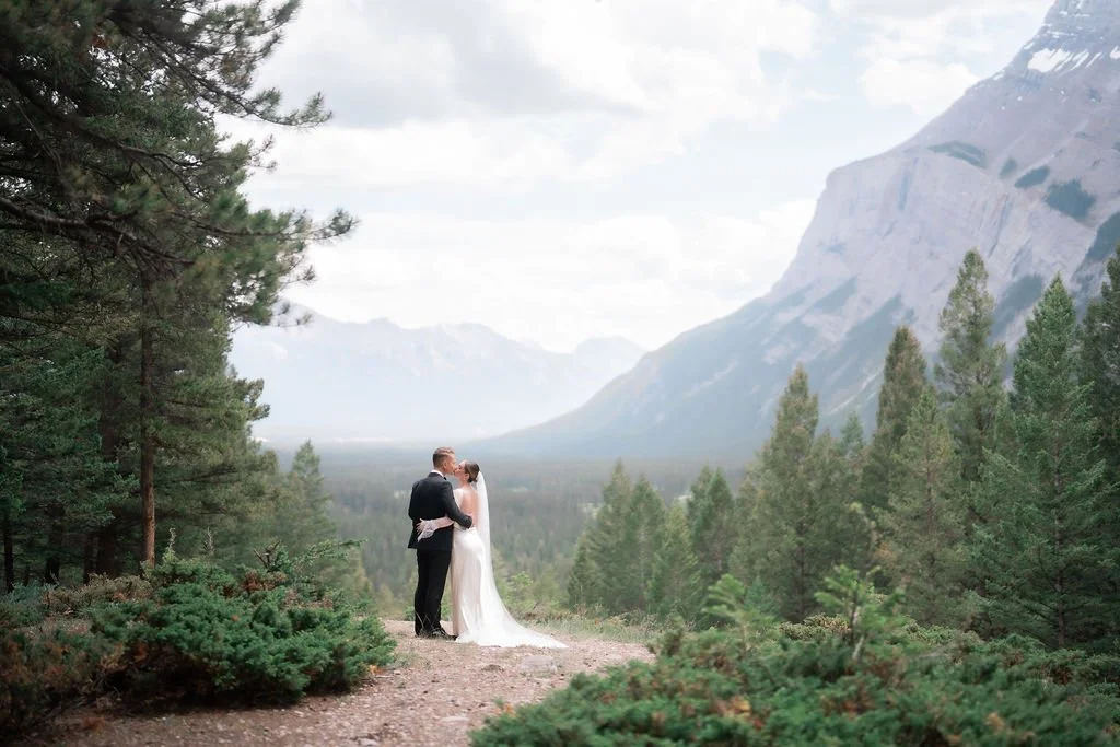 A bride and groom kissing in a forest with mountains in the background.