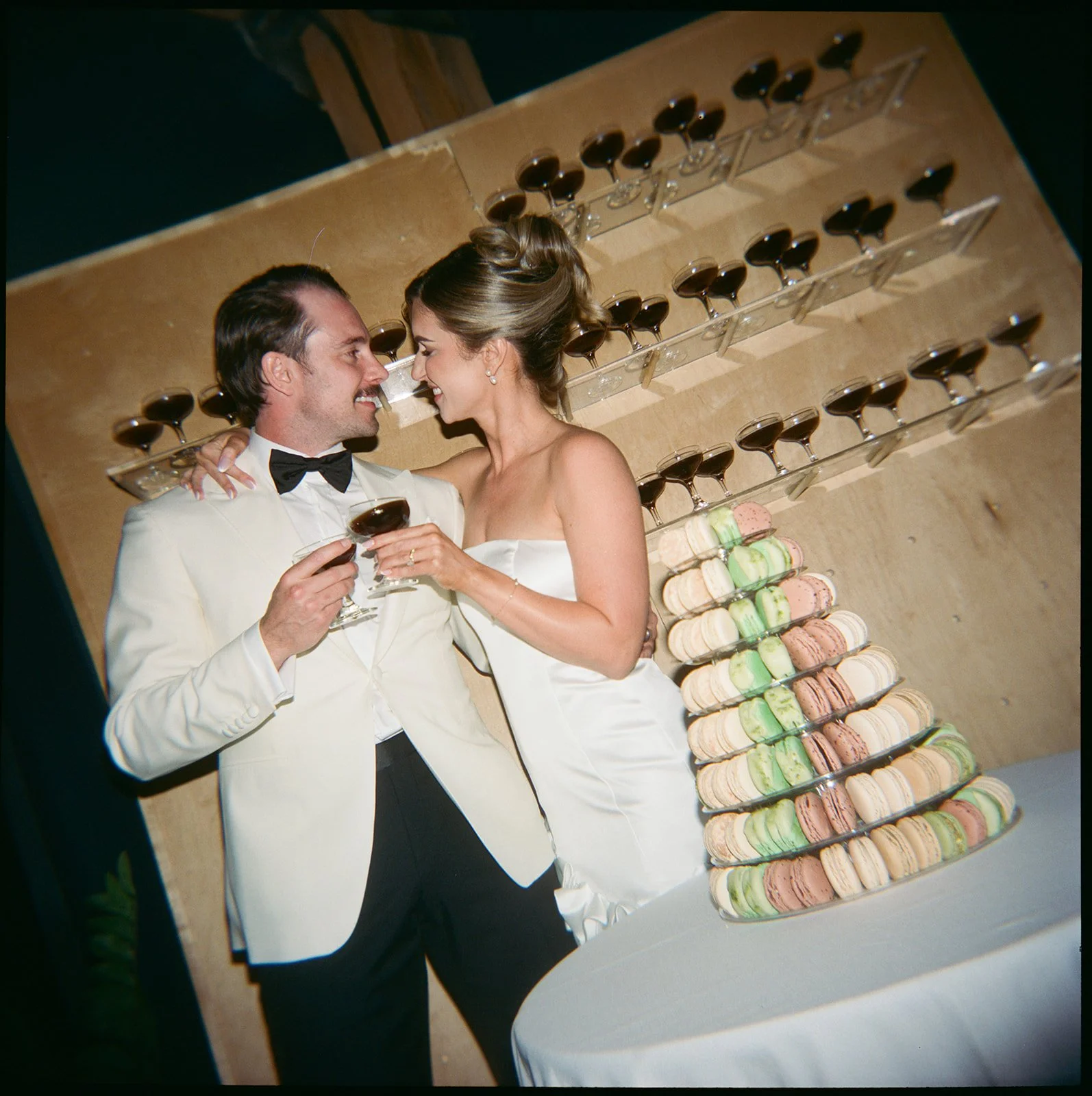 A newlywed couple in wedding attire sharing a toast at their wedding reception, with a colorful macaron display and multiple glasses of red wine in the background.
