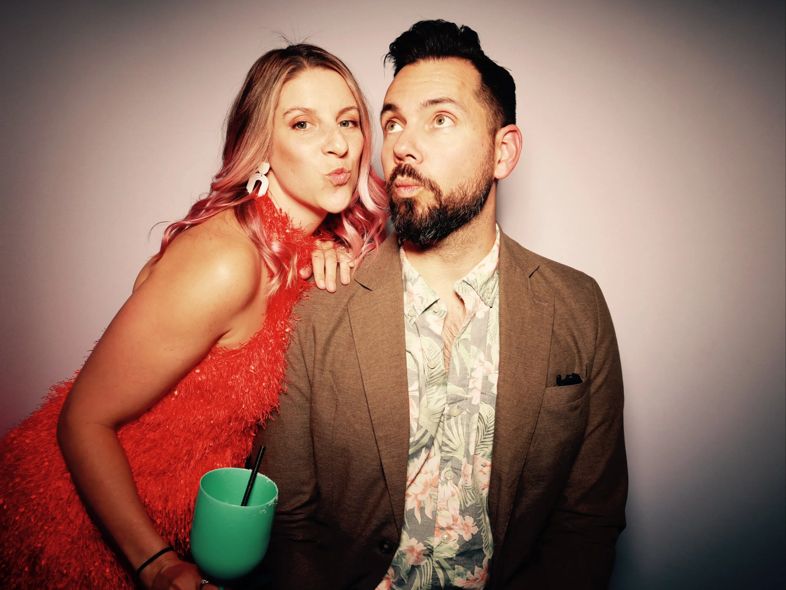 A woman with pink hair in a red dress leaning close to a man with dark hair and a beard, wearing a brown blazer and floral shirt, against a plain background.