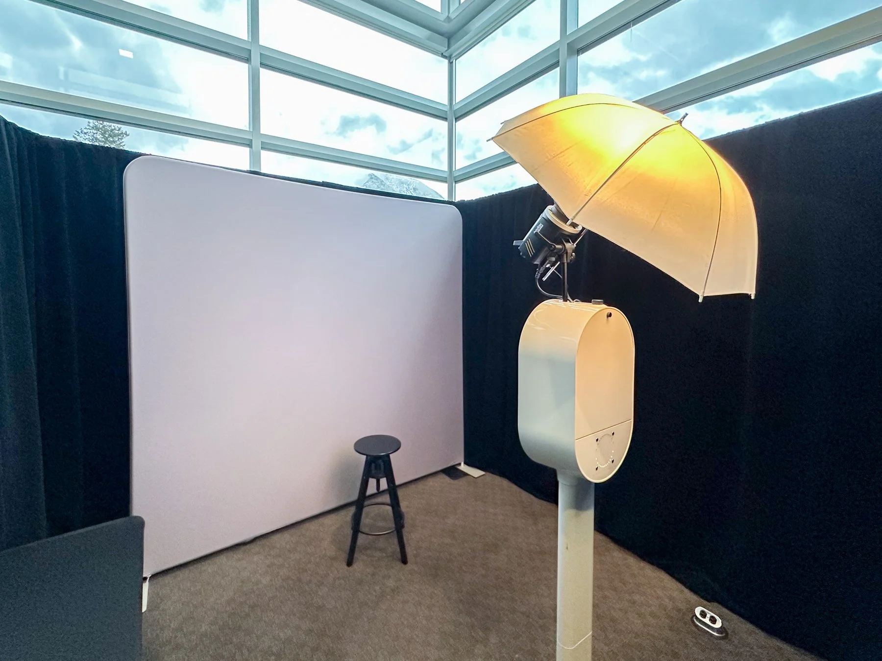 A photo of a photo booth with a large white curtain, a black stool, and a vintage yellow lampshade with a lamp underneath, set indoors near large windows.
