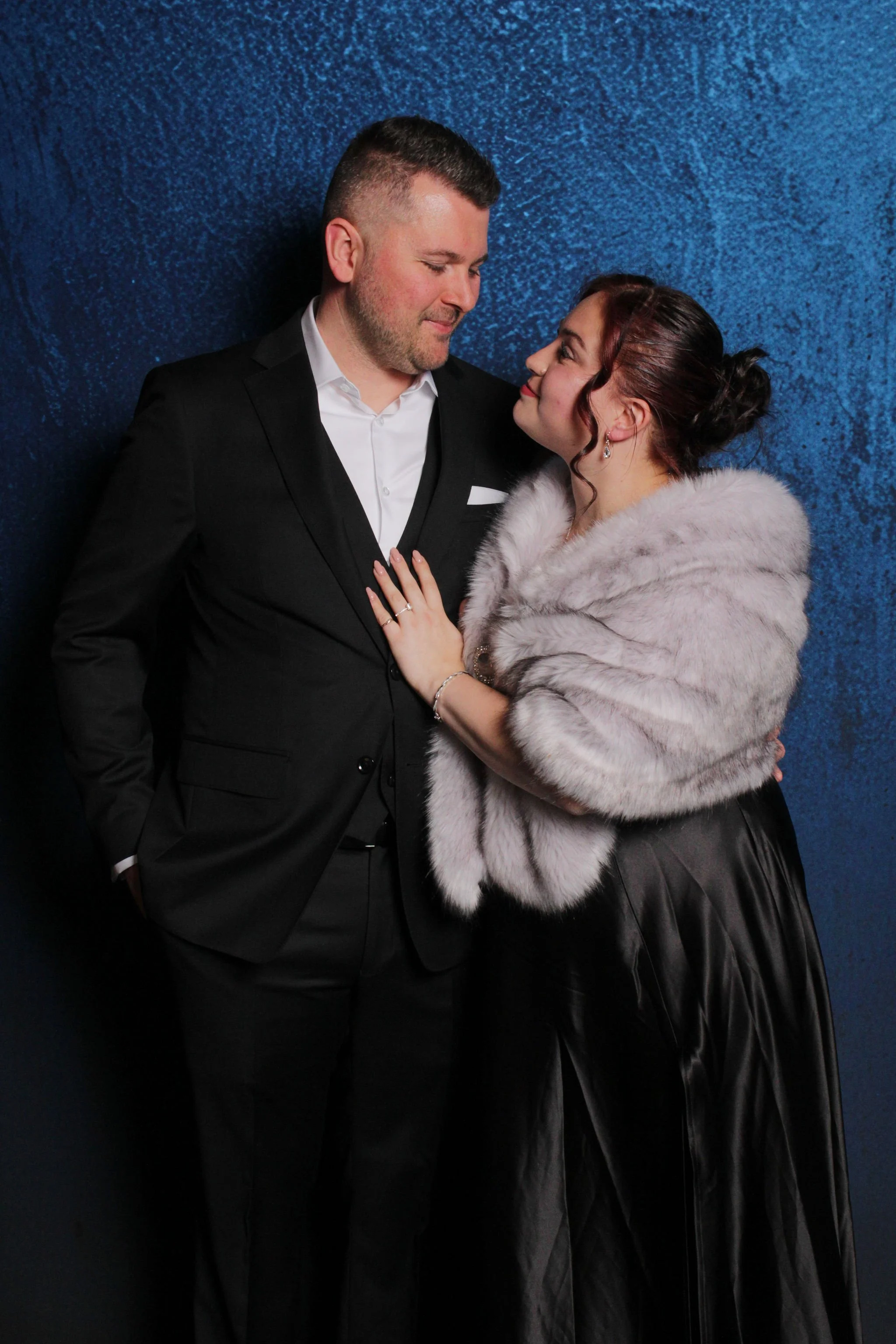 A man in a black suit and a woman in a gray fur stole and dark skirt are standing close, looking into each other's eyes against a blue textured background.