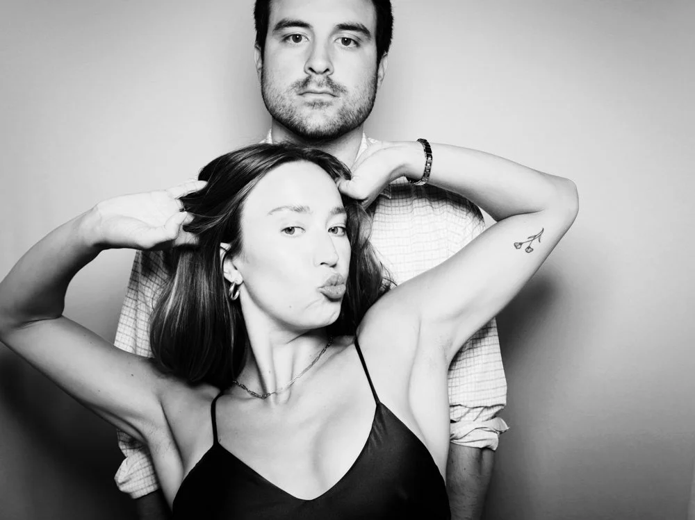 A black and white photo of a man standing behind a woman. The woman is making a duck face and flexing her arm, showing a tattoo of cherries. She is wearing a thin-strapped top and jewelry. The man has short hair, a beard, and is wearing a collared sh