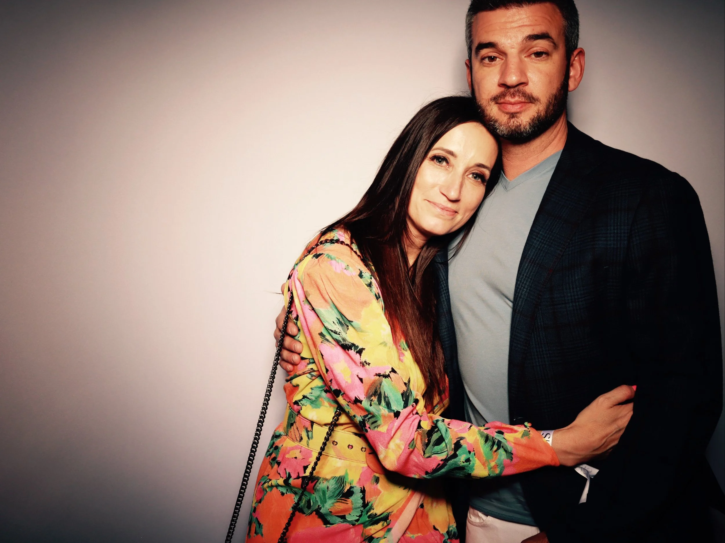 A man and a woman standing close together, smiling, against a plain background, with the woman resting her head on the man's chest.