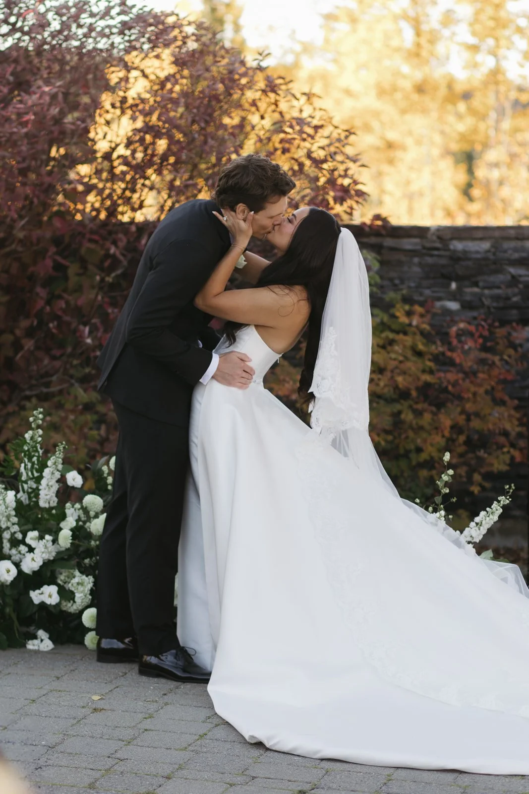 A bride and groom sharing a kiss outdoors during their wedding, with fall foliage in the background and flowers on the ground.