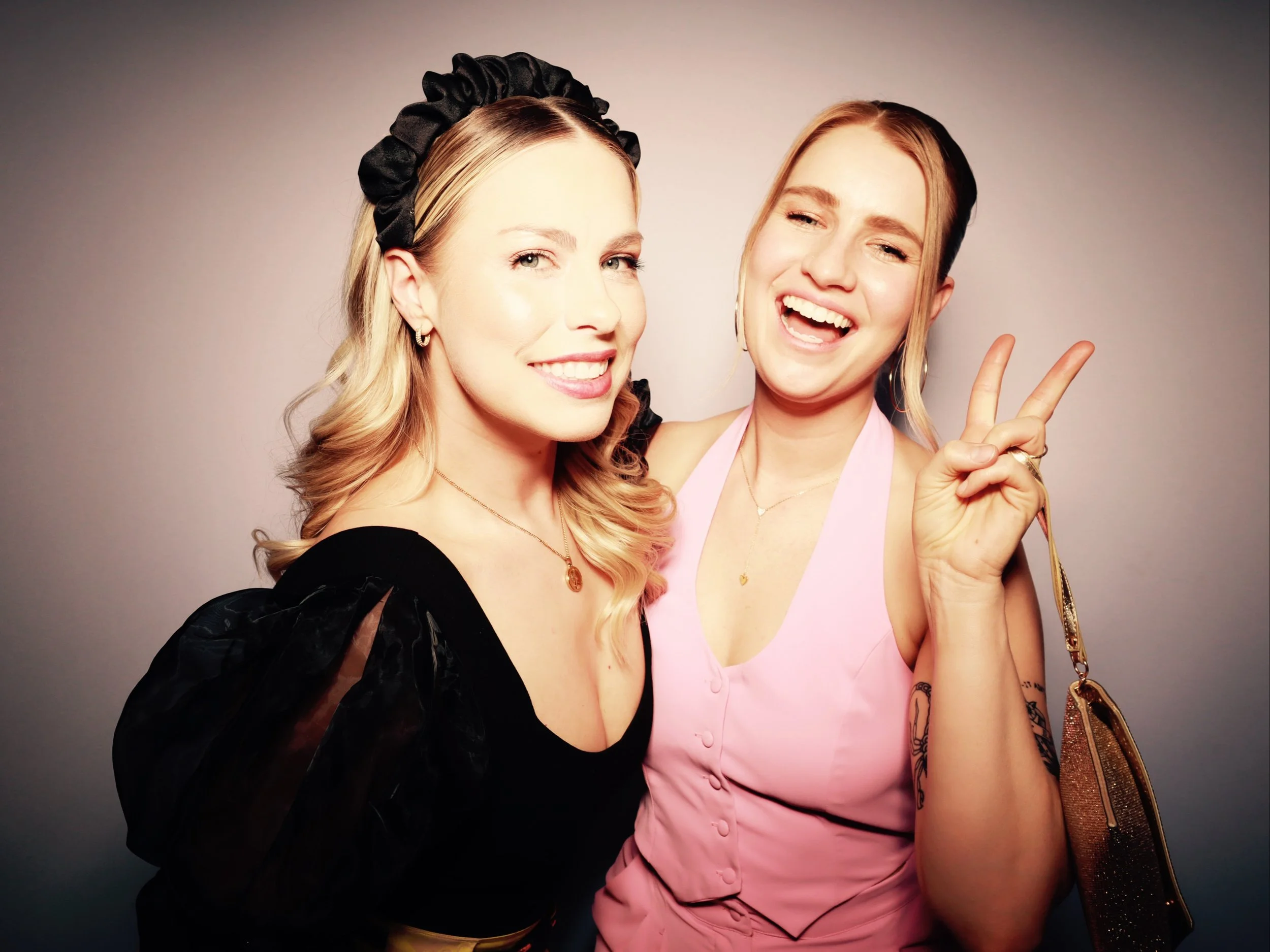 Two women smiling and posing for a photo, one making a peace sign with her hand, against a plain background.