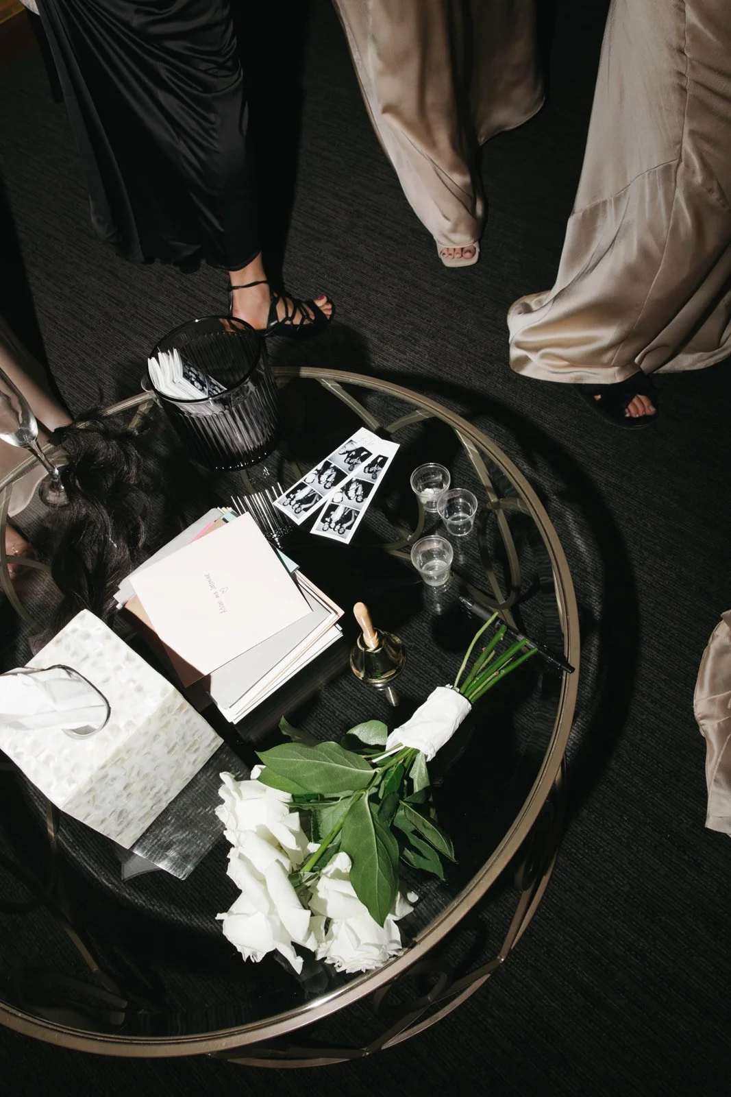 A glass-top table with a bouquet of white roses, photo booth strips, tracing paper, and various items; women in black and beige dresses are standing around.