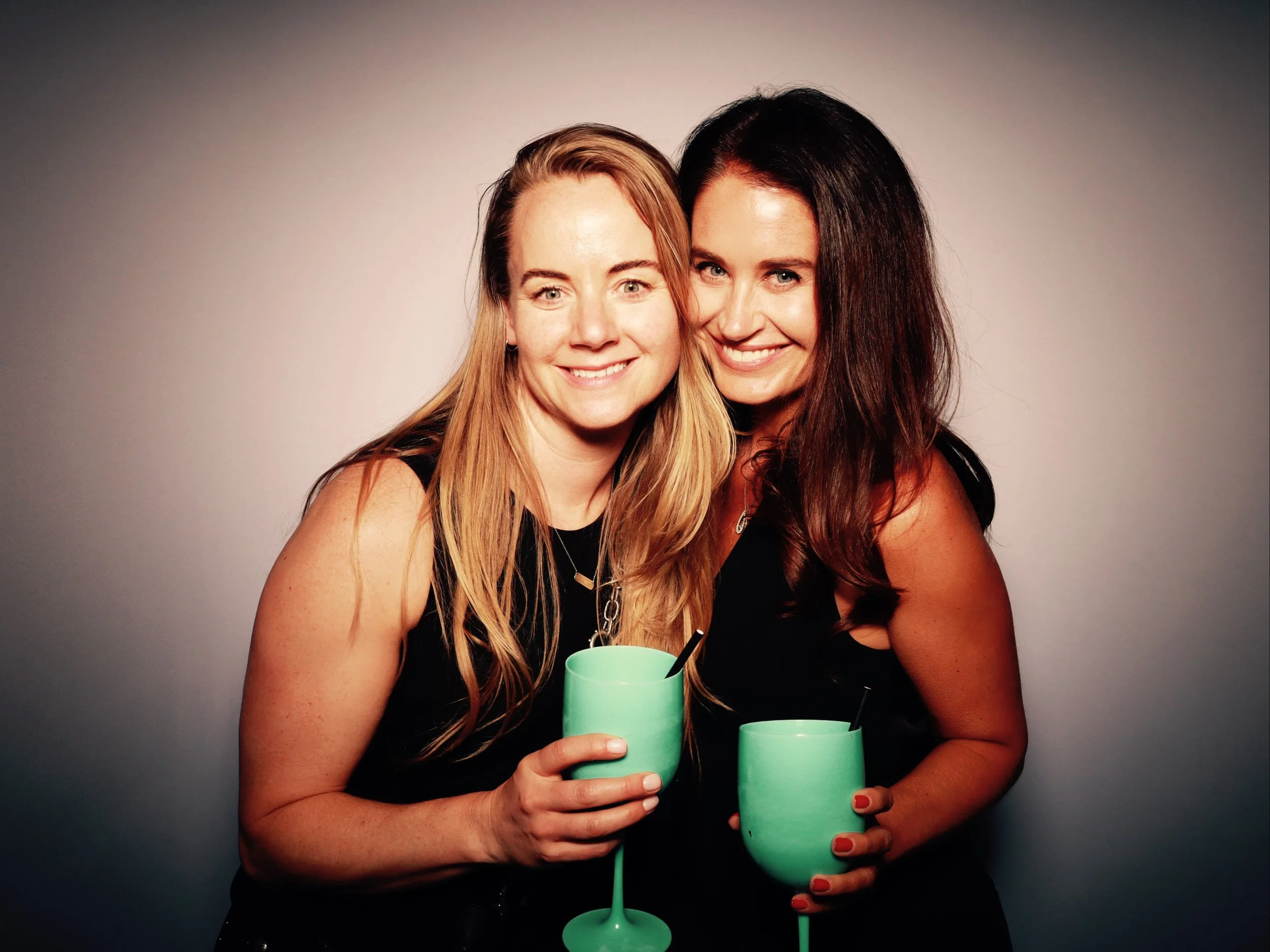 Two women smiling and holding green wine glasses with straws against a plain background.