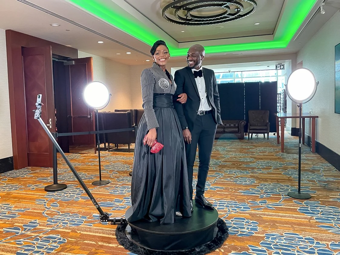 A couple dressed in formal attire with the woman in a black and white striped top and long black skirt, and the man in a black tuxedo with a bow tie, posing together on a circular platform in a well-lit event venue with green lighting accents on the 