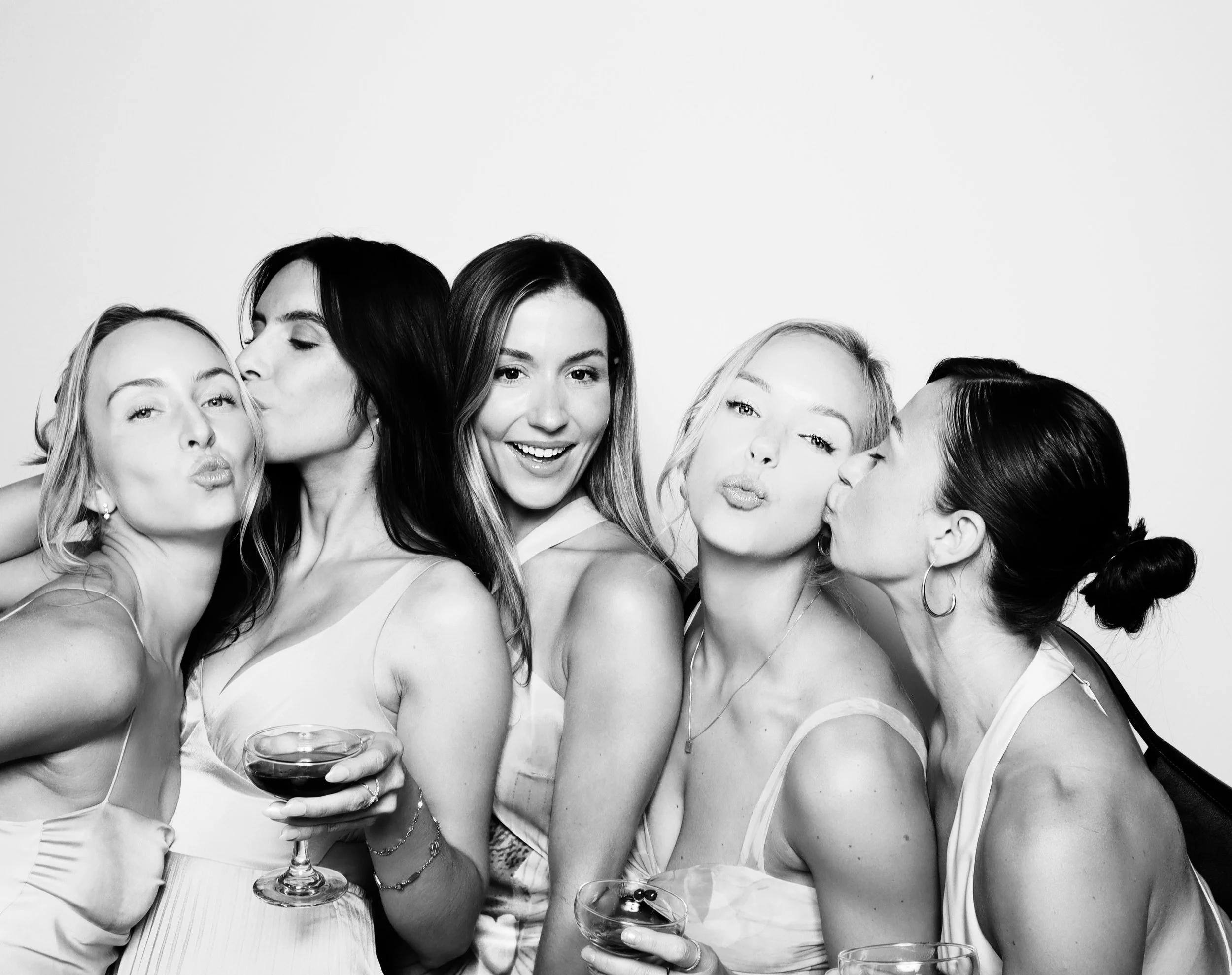 Group of five women enjoying a party, with some kissing and blowing kisses, one woman holding a glass of wine, all smiling, in black and white.