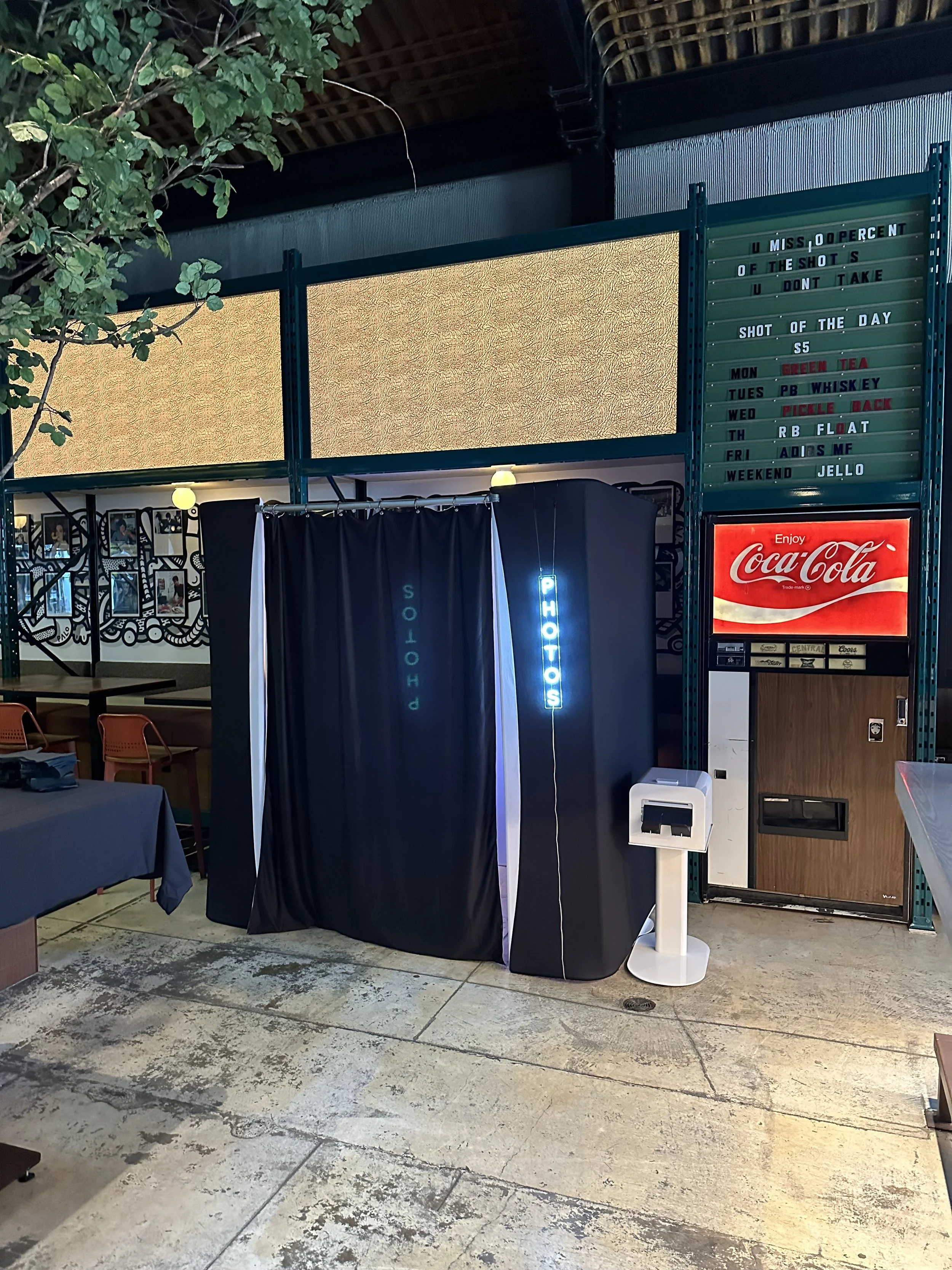 Photo of a photo booth with black curtains labeled 'PHOTOS' in neon blue, situated outdoors with tables and chairs nearby, under a partially visible leafy tree and a high ceiling.