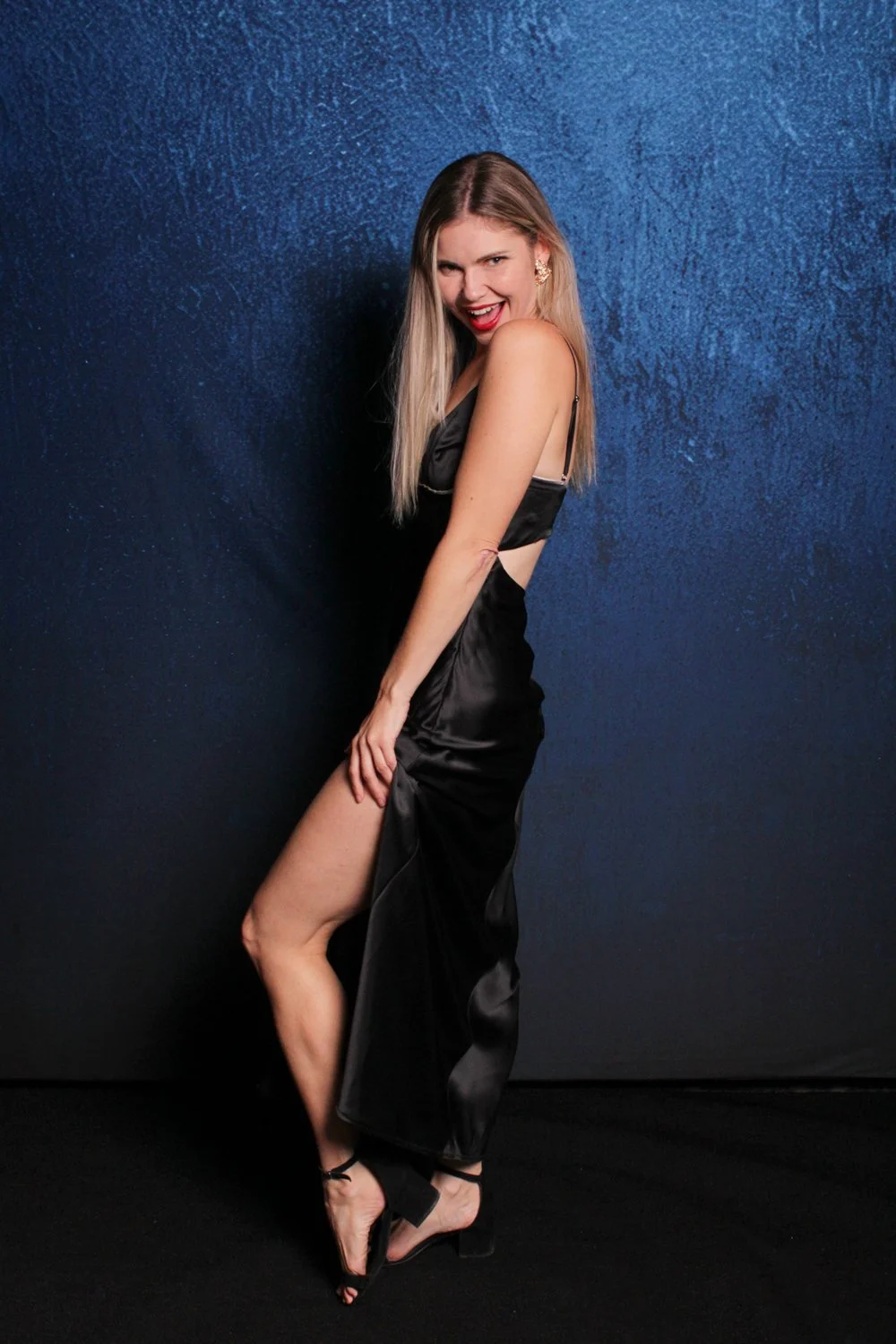 A woman in a black satin dress with a high slit, black high heels, and gold earrings, posing confidently with a playful expression against a textured dark blue background.