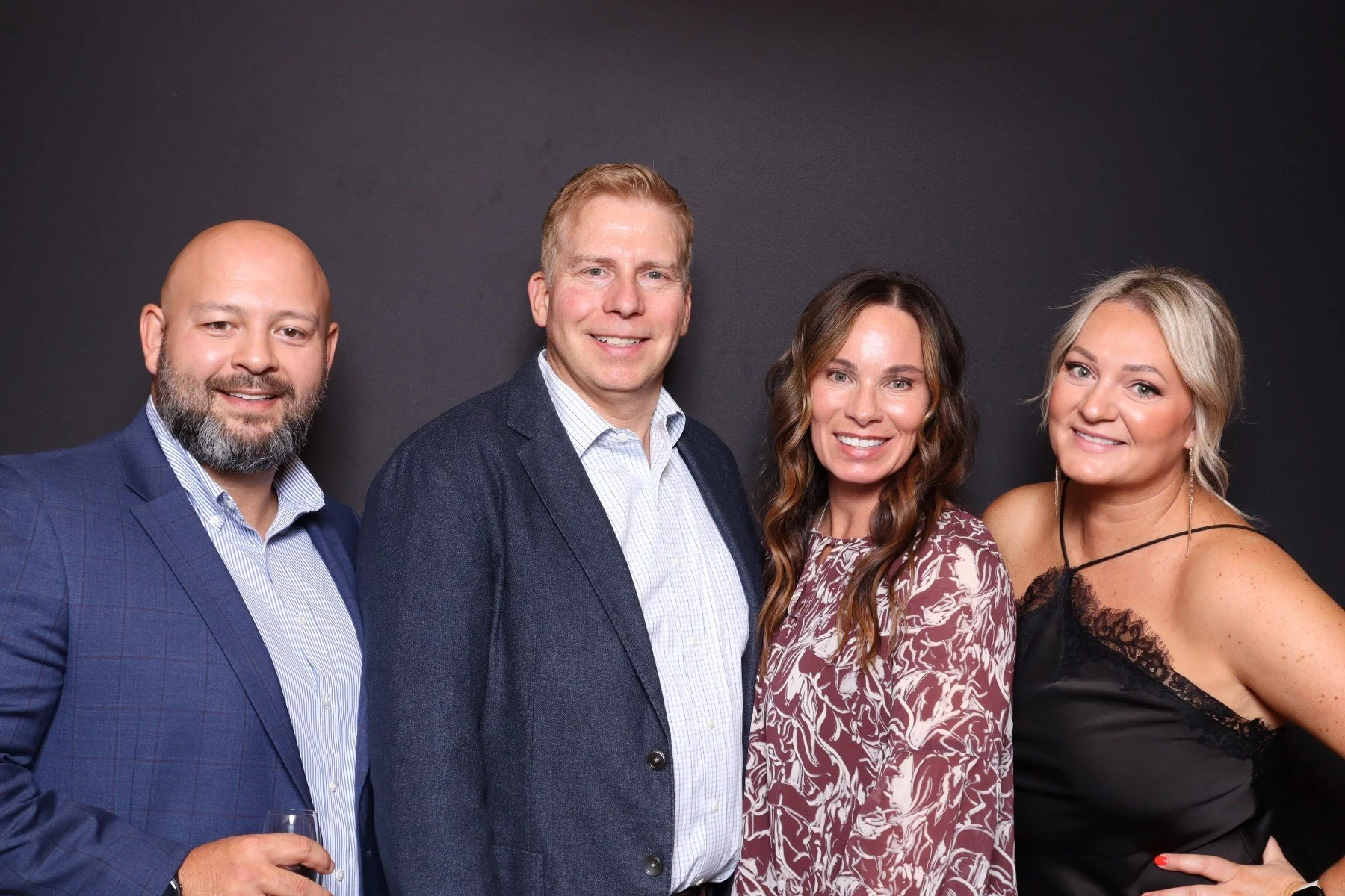 Four adults in formal attire, smiling and posing in front of a plain dark background. The man on the left has a beard and is holding a glass of drink. The man next to him has short blond hair and is wearing a navy blazer. The woman next to him has lo