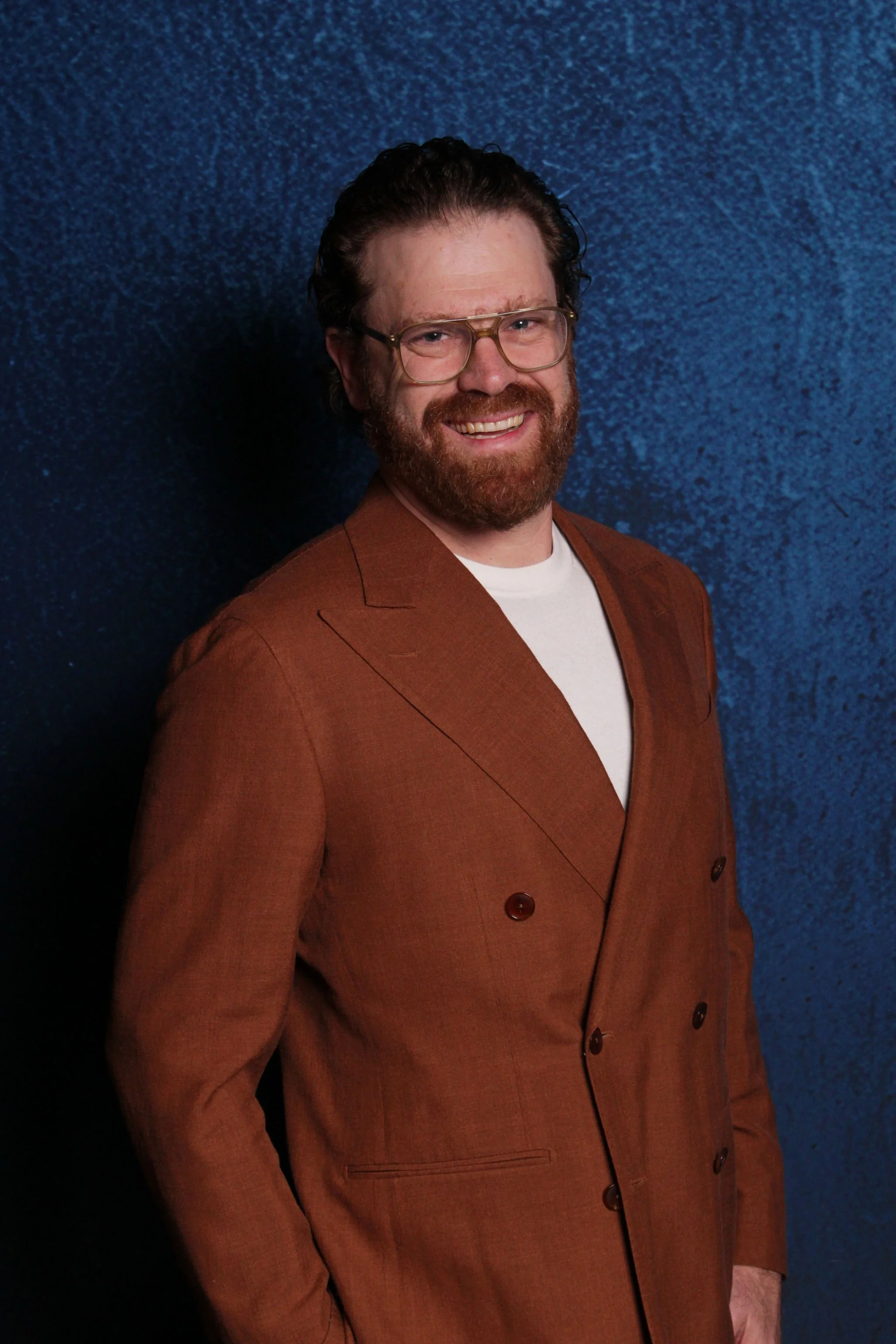 A man with glasses, a beard, and curly hair smiling in front of a blue textured background, wearing a brown double-breasted blazer and a white shirt.