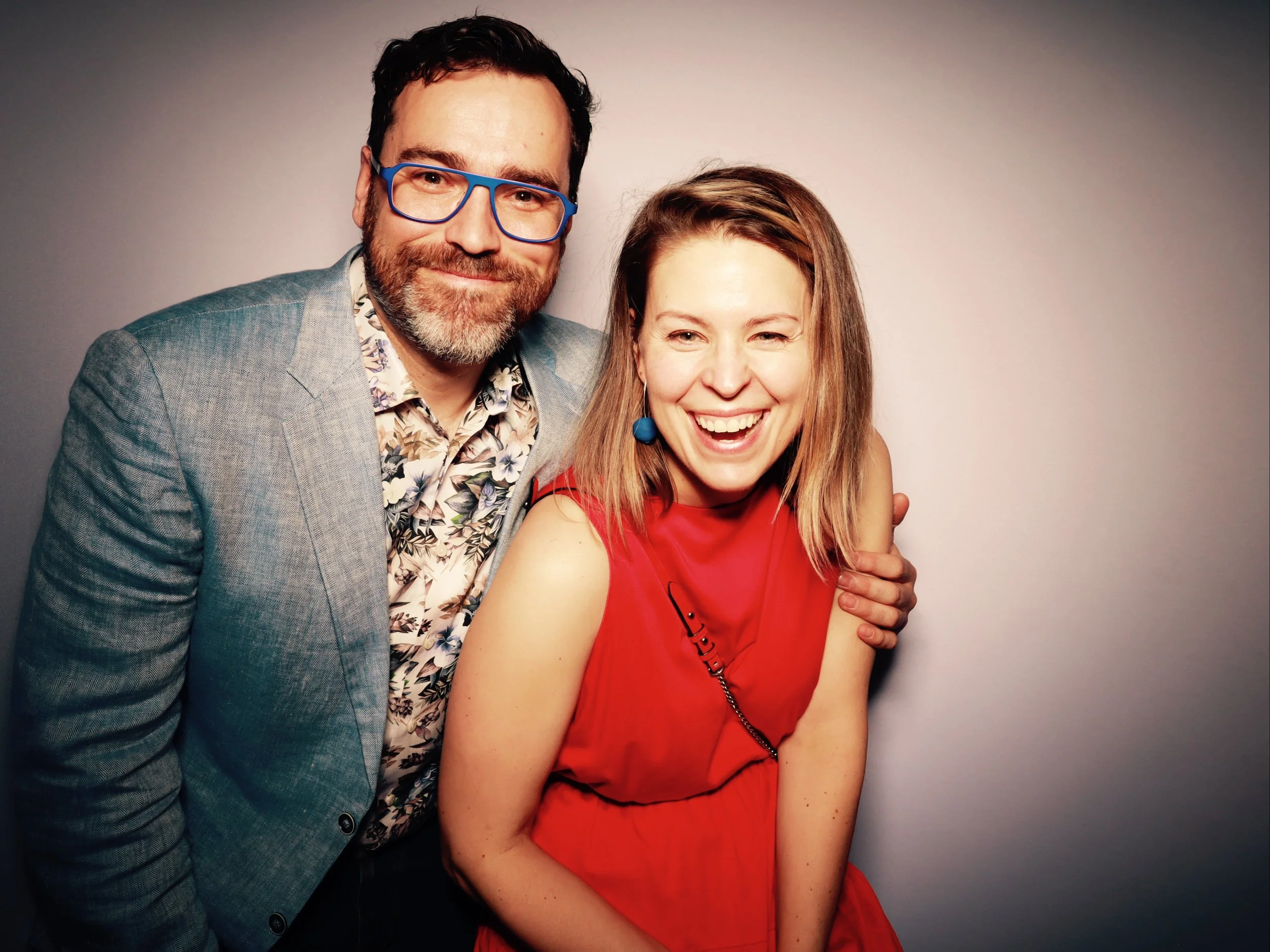 A man with a beard and blue glasses in a gray blazer, and a woman with blonde hair in a red sleeveless dress, both smiling and laughing in front of a plain background.