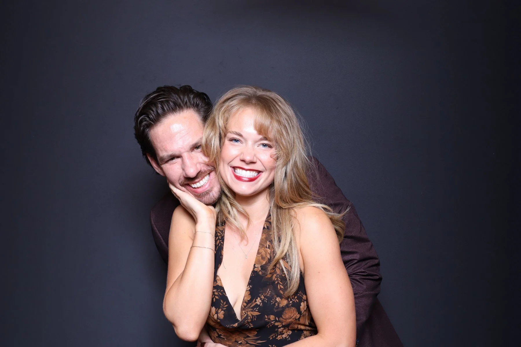 A happy couple smiling and posing together in front of a dark background. The woman has long blonde hair and is wearing a sleeveless floral dress, while the man has dark hair and is wearing a dark suit.