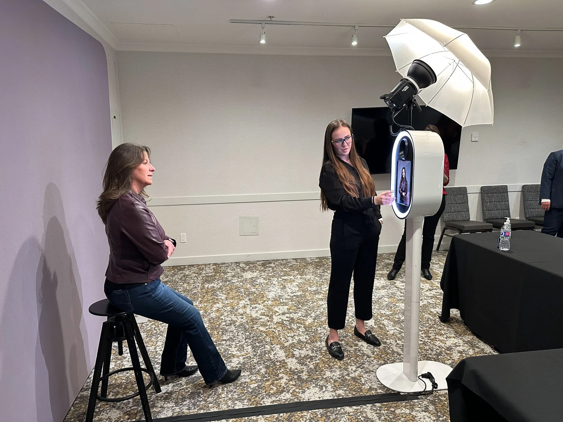 A woman sits on a stool in front of a light purple wall while another woman stands in front of a photo booth with a large umbrella and camera, preparing to take her picture. There are chairs and a table with a water bottle in the background.
