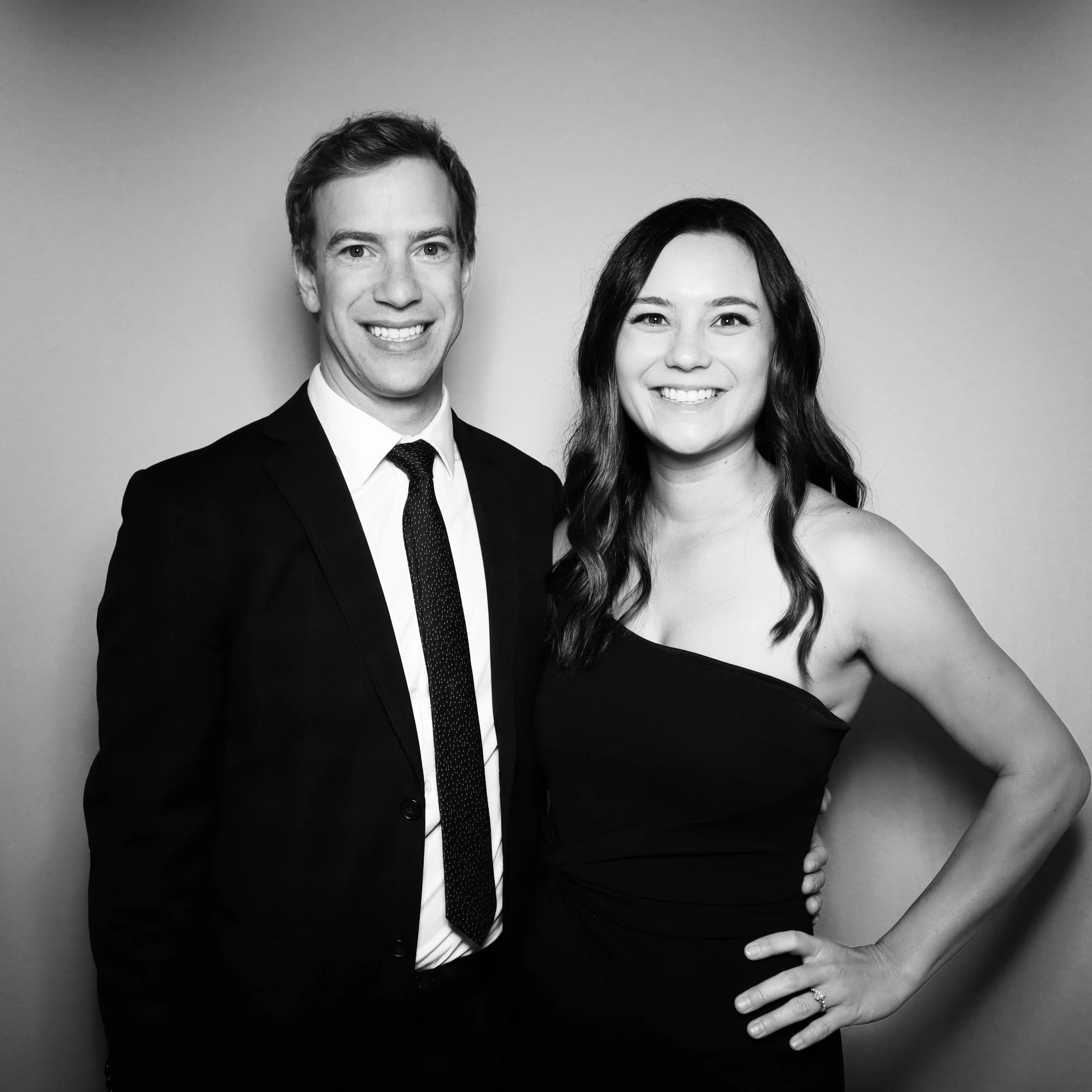 Black and white photo of a man in a suit and woman in a strapless dress smiling and standing close together against a plain background.