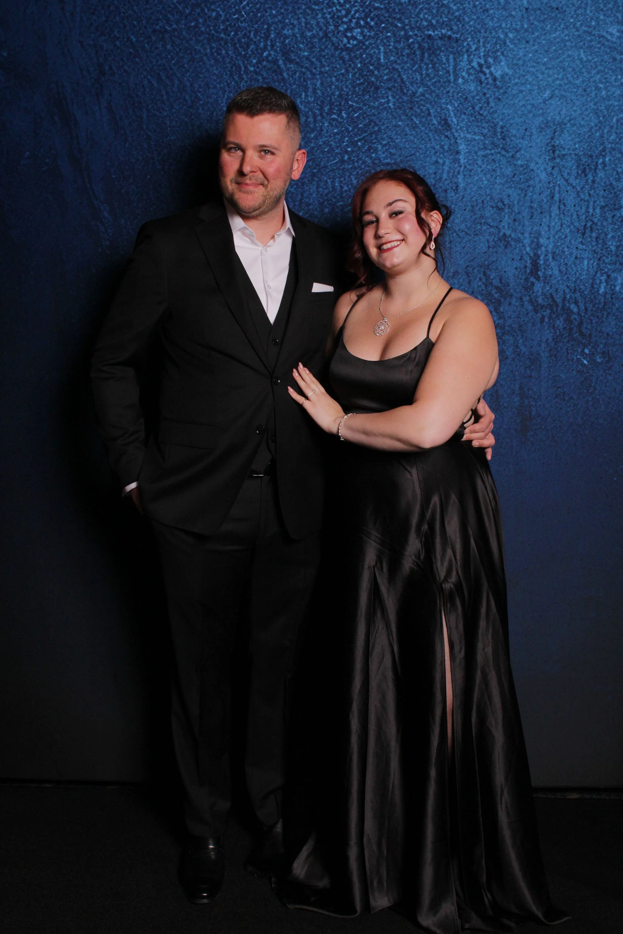 A man in a black suit and white shirt standing next to a woman in a black satin evening gown, both smiling, against a blue textured wall.