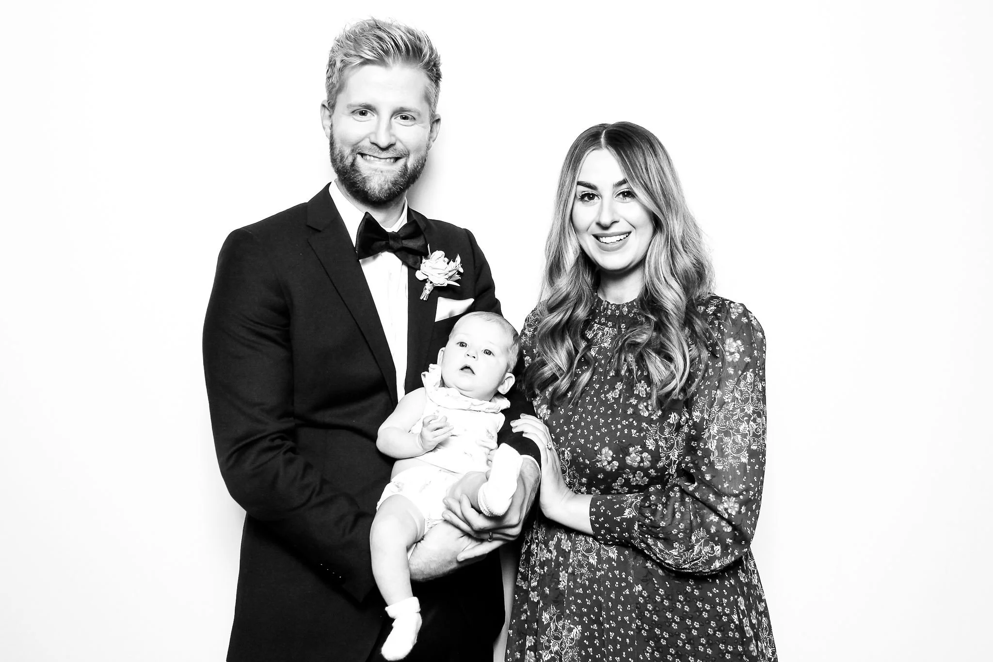 A black and white photo of a family with a man in a tuxedo, woman in a floral dress, and a baby.