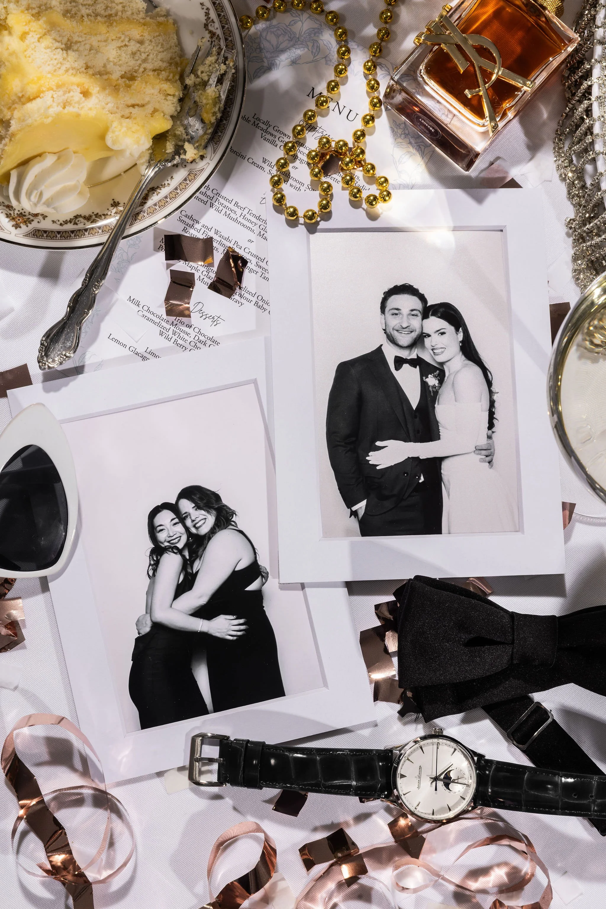 Decorative table top with framed black-and-white wedding photos, a wristwatch, a wedding dress bow tie, a beaded necklace, a perfume bottle, a plate of cake, chocolates, and scattered ribbons.
