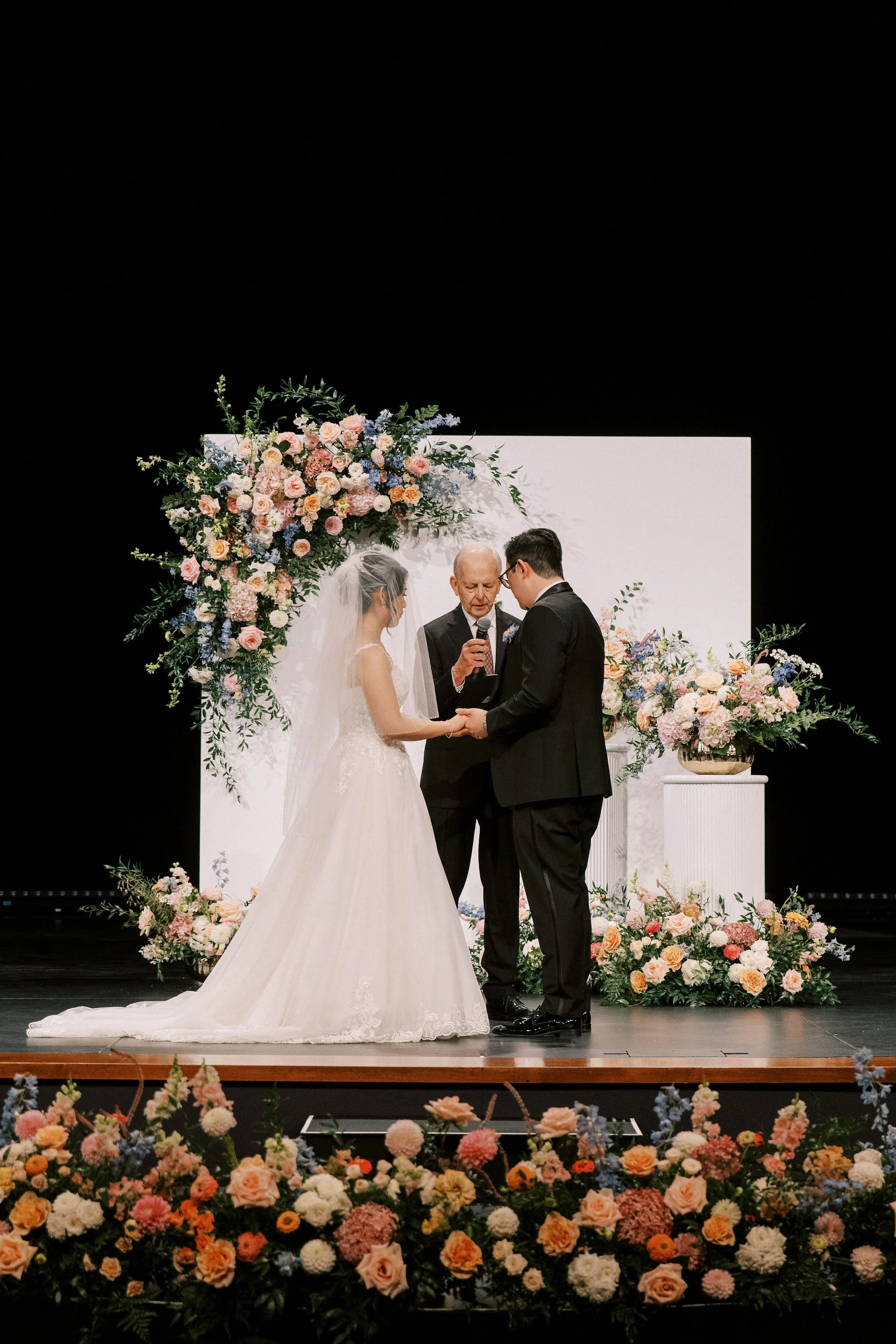 A wedding ceremony with a bride and groom holding hands, standing before an officiant, surrounded by floral arrangements on stage.
