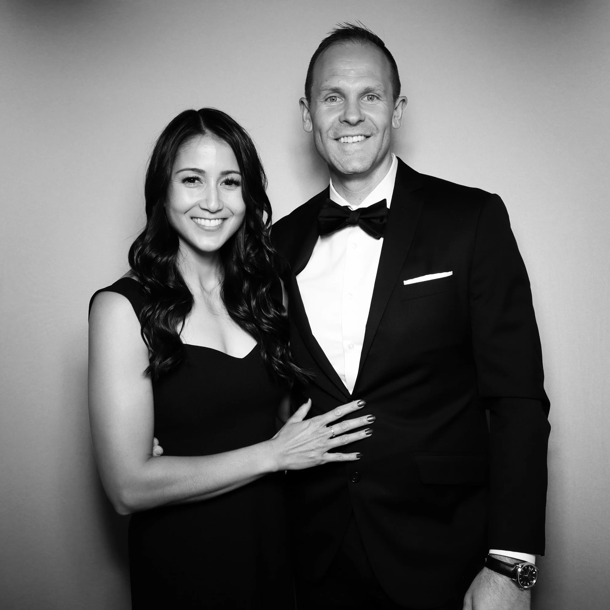 A smiling couple dressed in formalwear, the woman in a sleeveless black dress and the man in a tuxedo with a bow tie, standing together in front of a plain background.