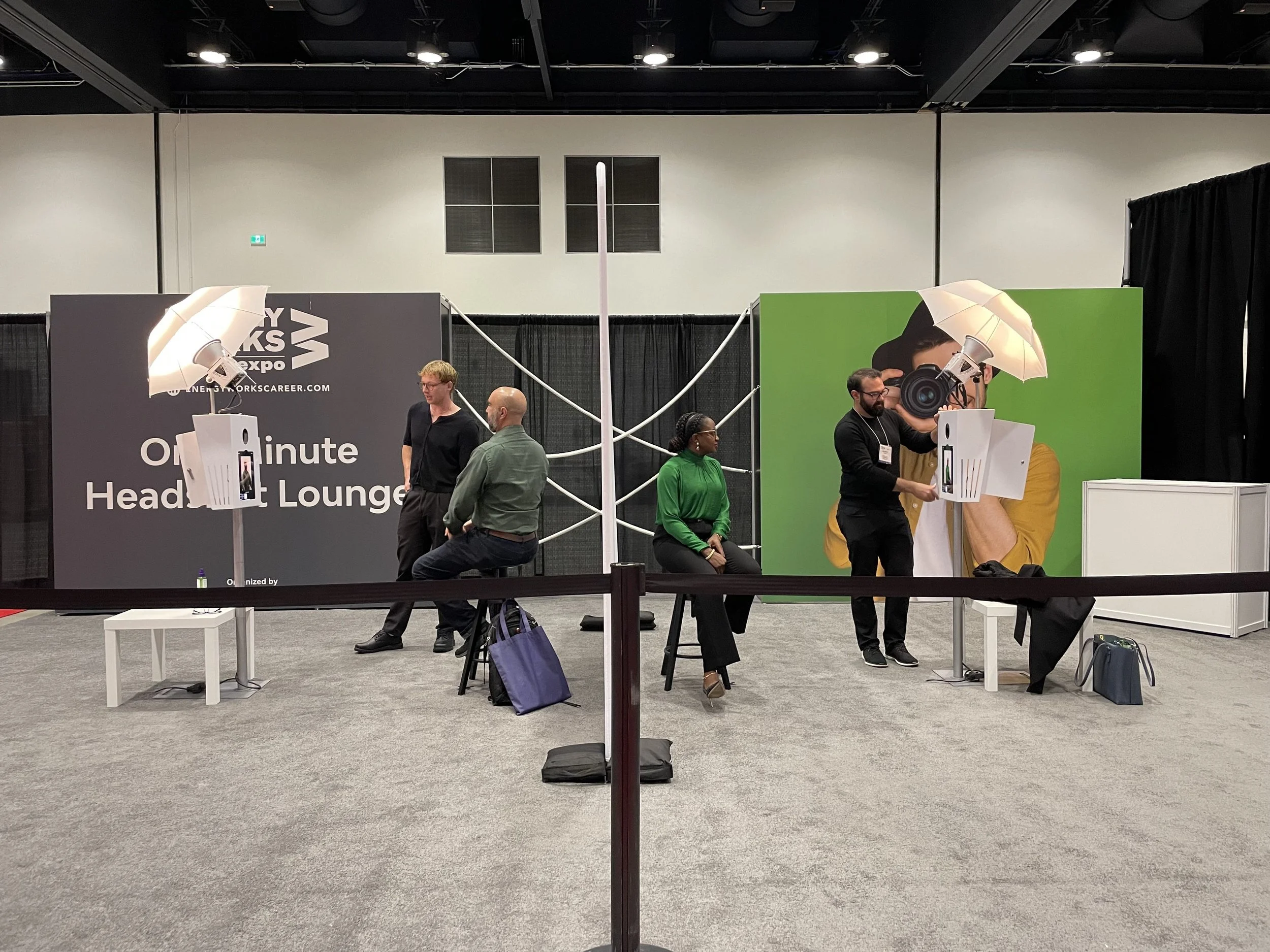 A photo of a photo booth setup at a convention, featuring large printed backdrops with people sitting and standing, overhead studio lights, and a person taking a digital photo of a woman with a camera icon on a green background.