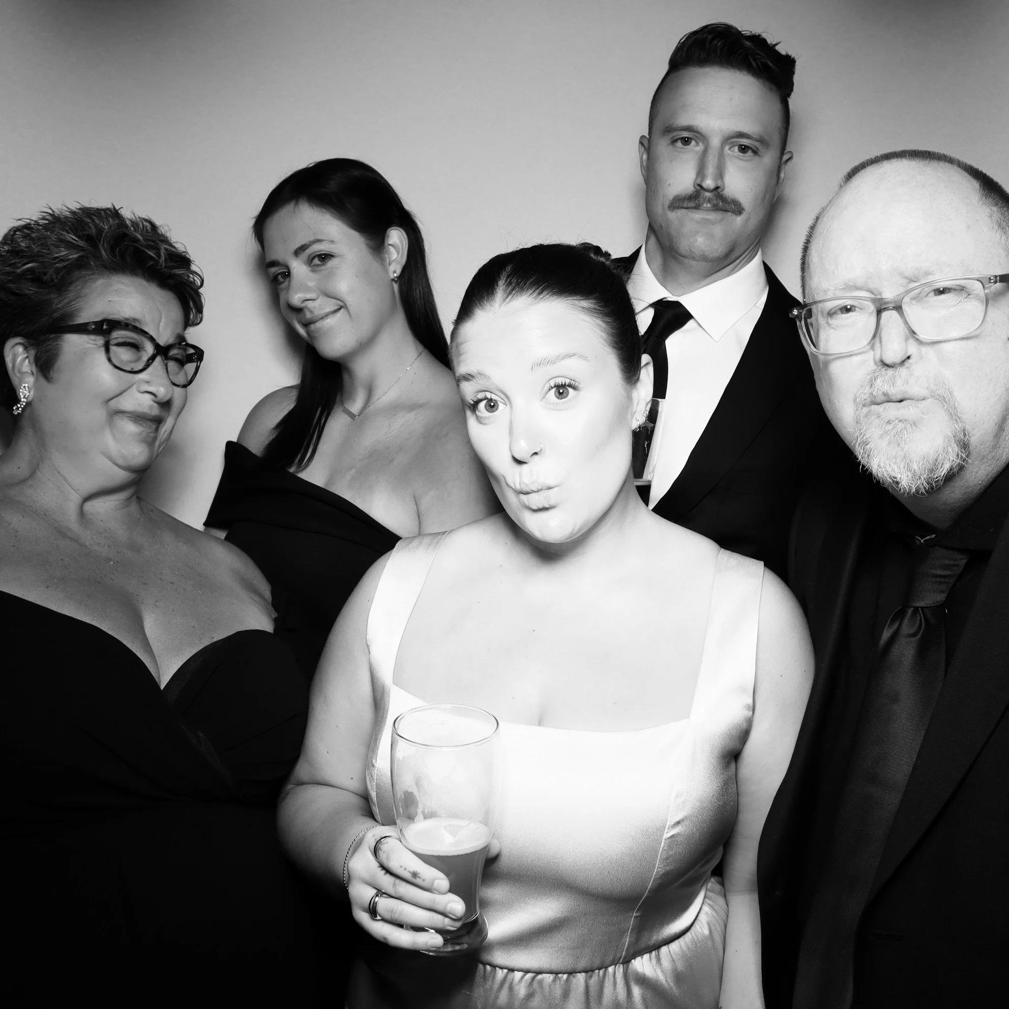 Group of five adults at a formal event, with one woman holding a glass, dressed in elegant attire, posing for a photo in black and white.