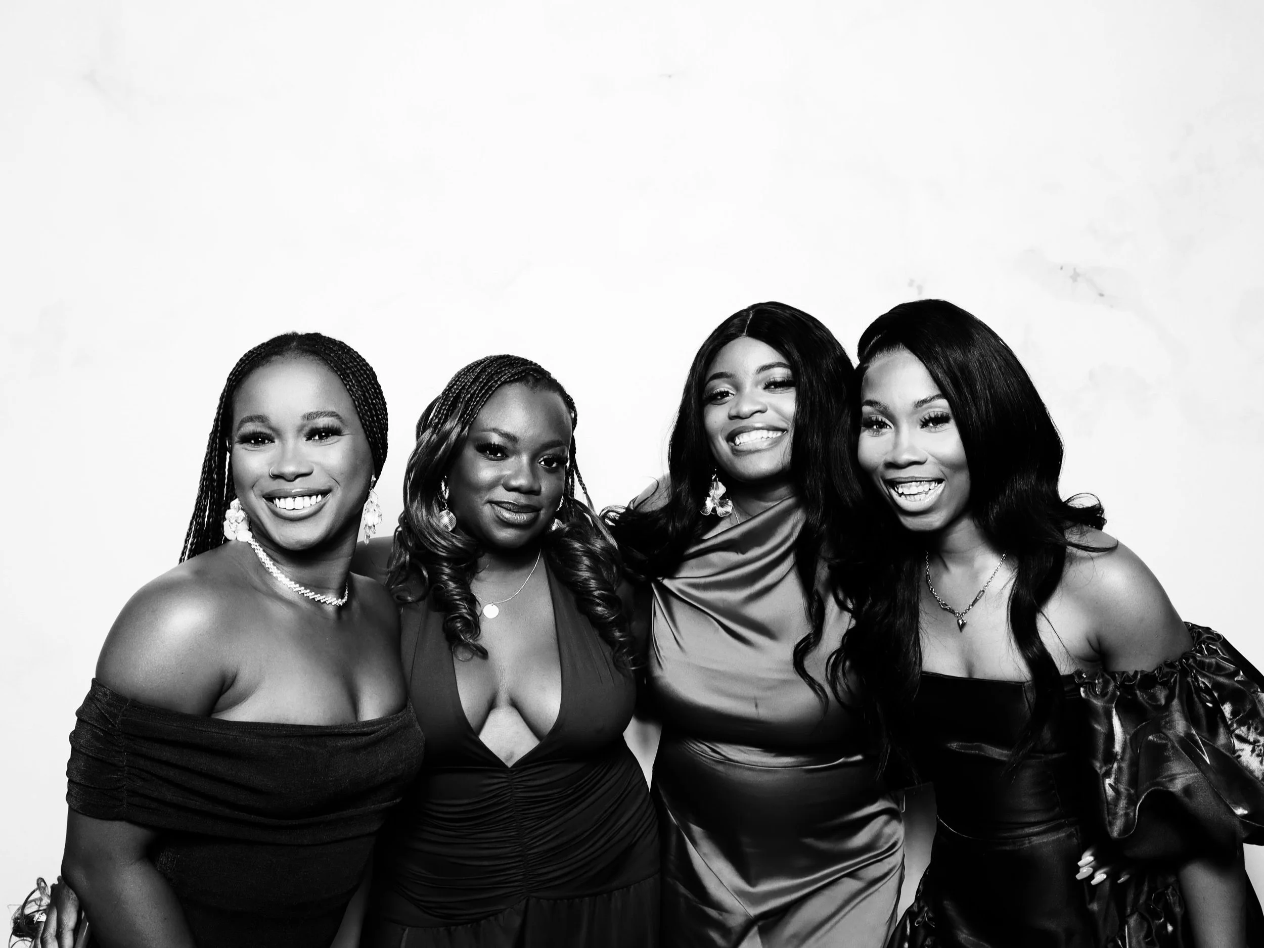 Four women dressed elegantly stand closely together, smiling at the camera against a plain background.