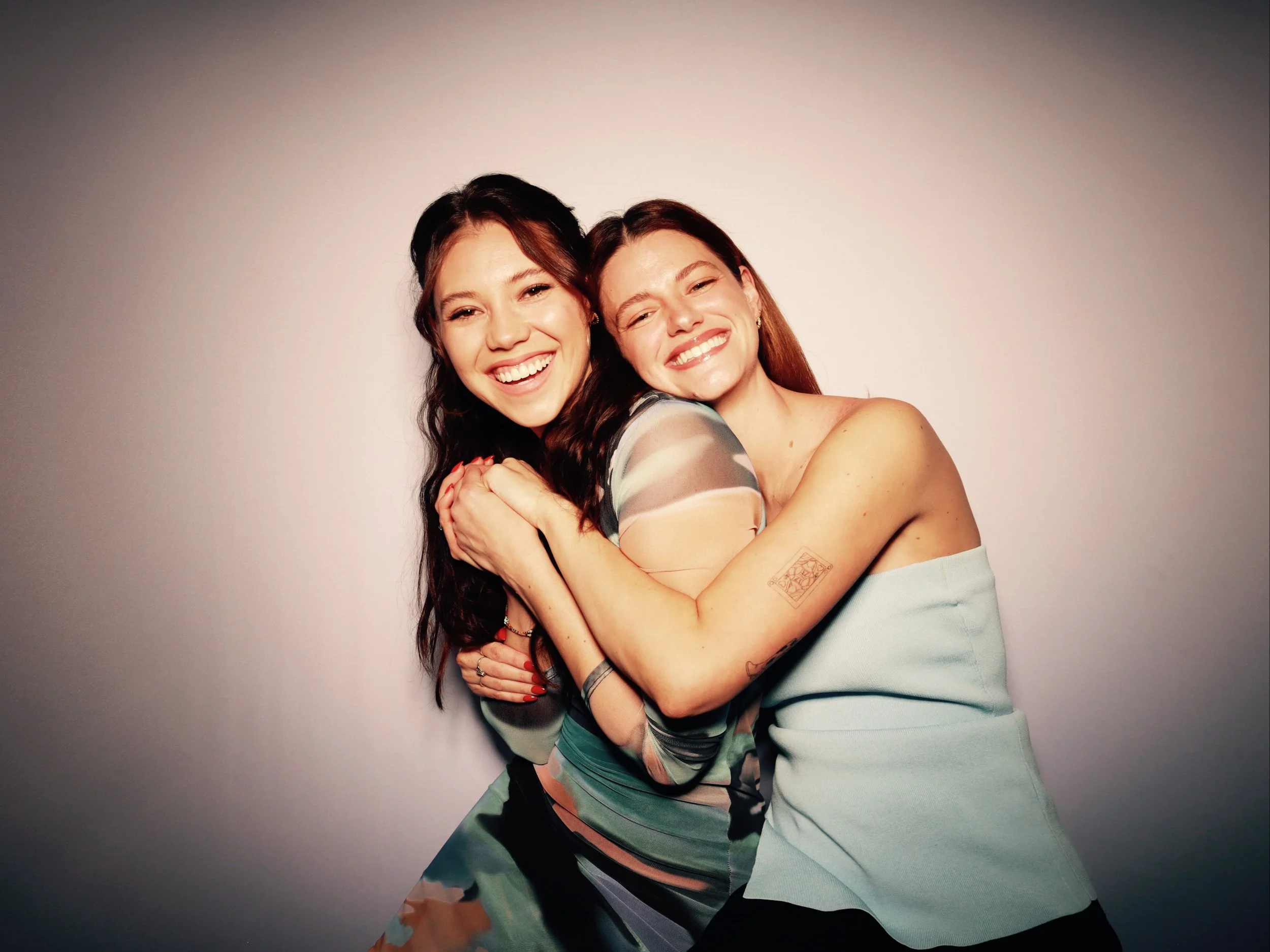 Two women with brown and red hair smiling and hugging each other against a plain light-colored background.