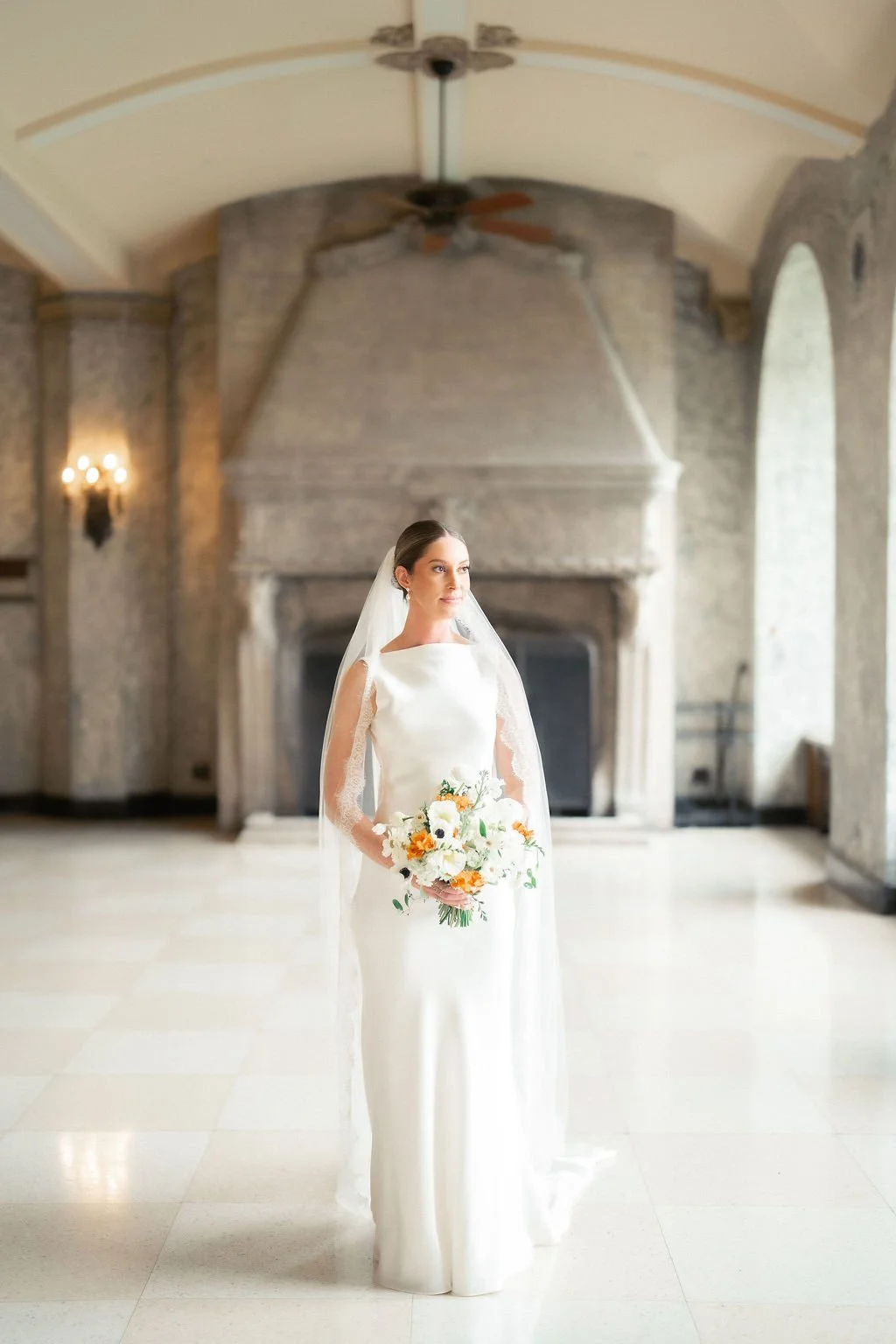 A bride in a white wedding gown holding a bouquet of white and orange flowers, standing in a spacious room with stone walls and a fireplace.