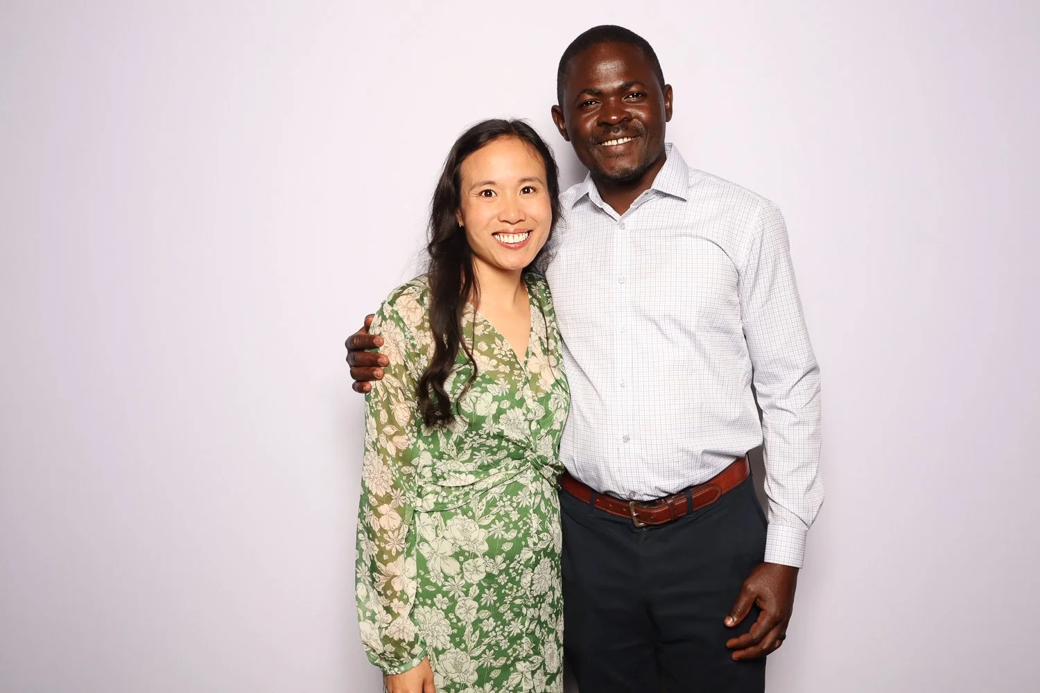 A smiling woman and man standing close together, with the woman's arm around the man's back and the man's arm around the woman's shoulder, against a plain light background.