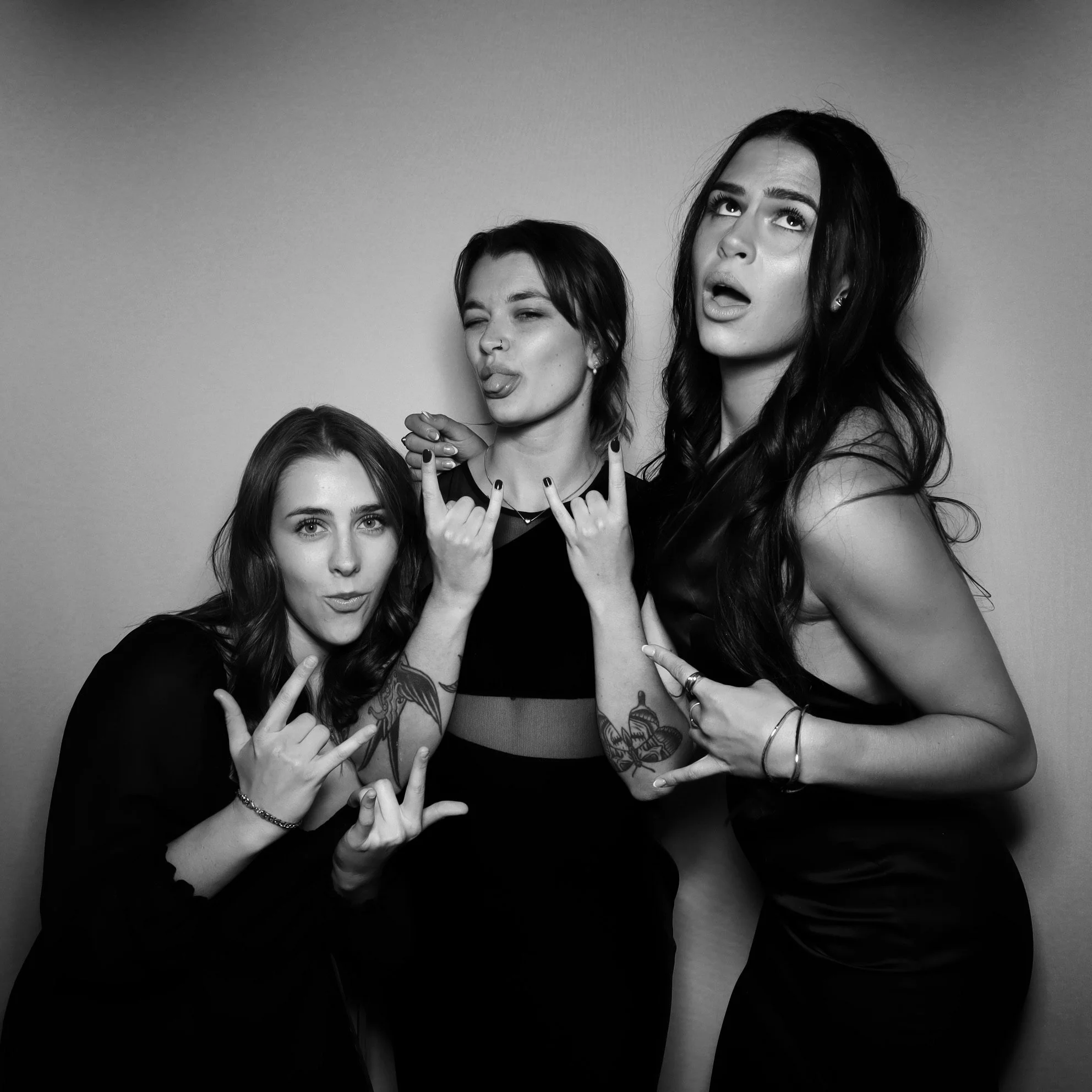 Three women posing and making rock and roll hand gestures, with their tongues out, in a black and white photo.