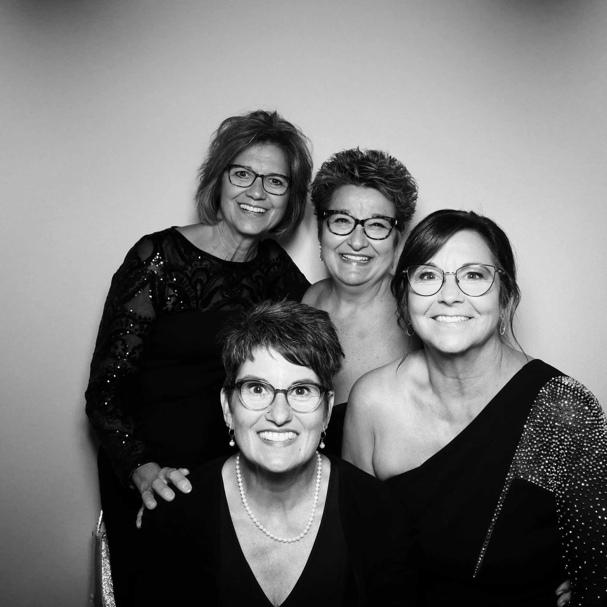 Black and white photo of five smiling women wearing glasses, dressed in elegant attire, posing against a plain background.
