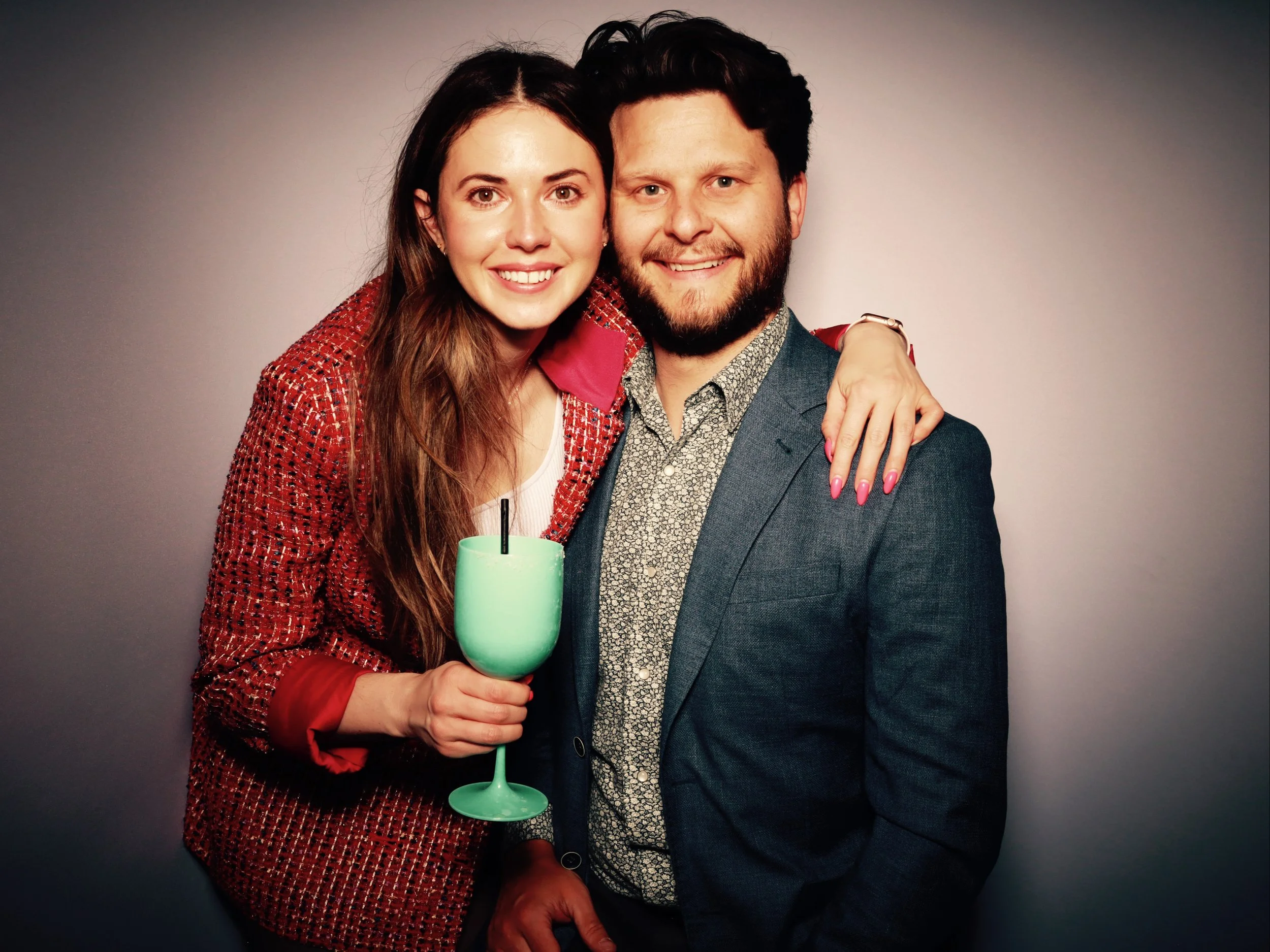 A woman with long brown hair wearing a red patterned blazer and white top, smiling and holding a turquoise cocktail with a straw, standing next to a man with dark hair and beard wearing a dark blazer and patterned shirt, both smiling and posing toget