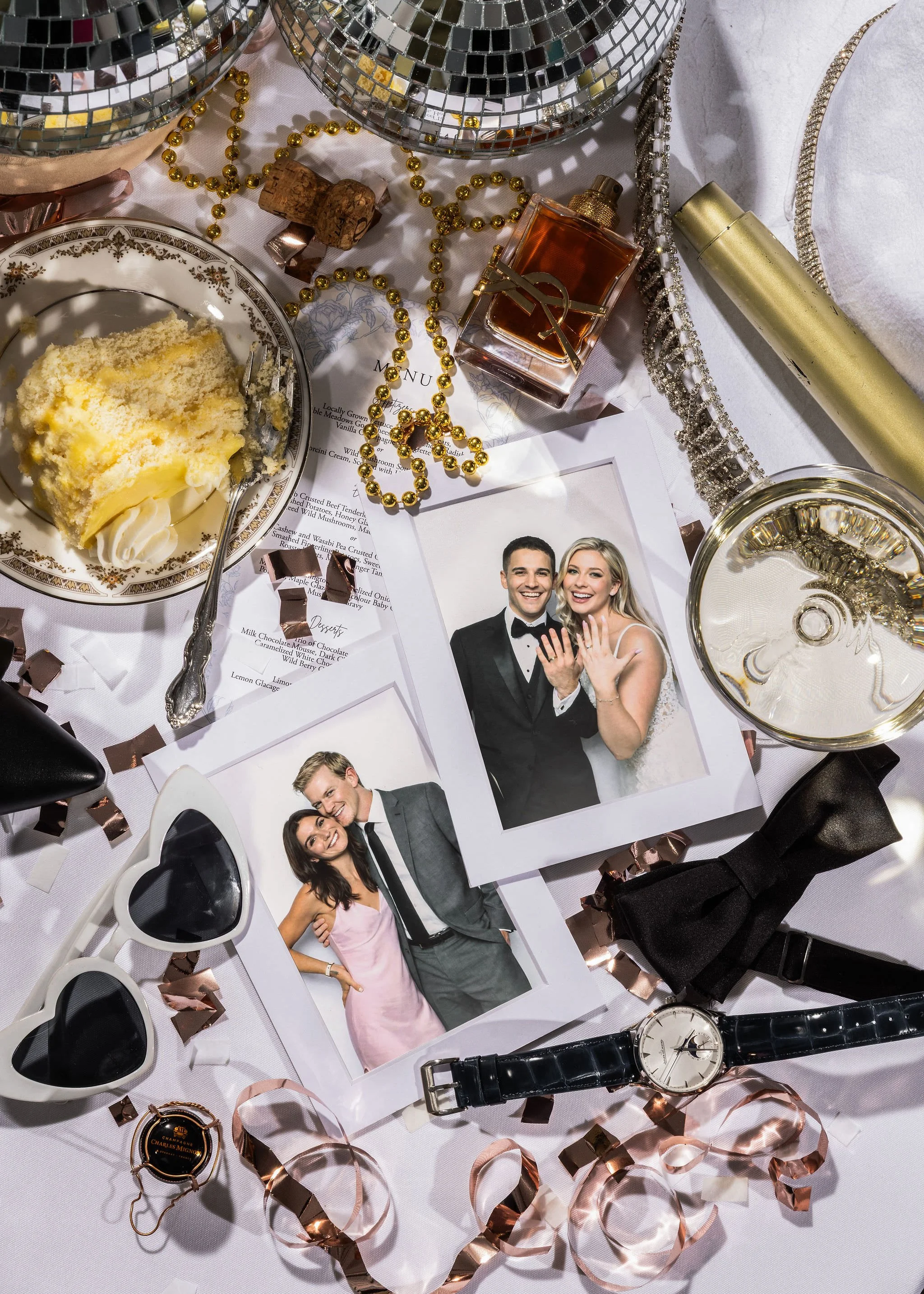A cluttered table with wedding photos, a slice of cake, sunglasses, a watch, a bow tie, a perfume bottle, and decorative items such as ribbons, beads, and a disco ball reflection.