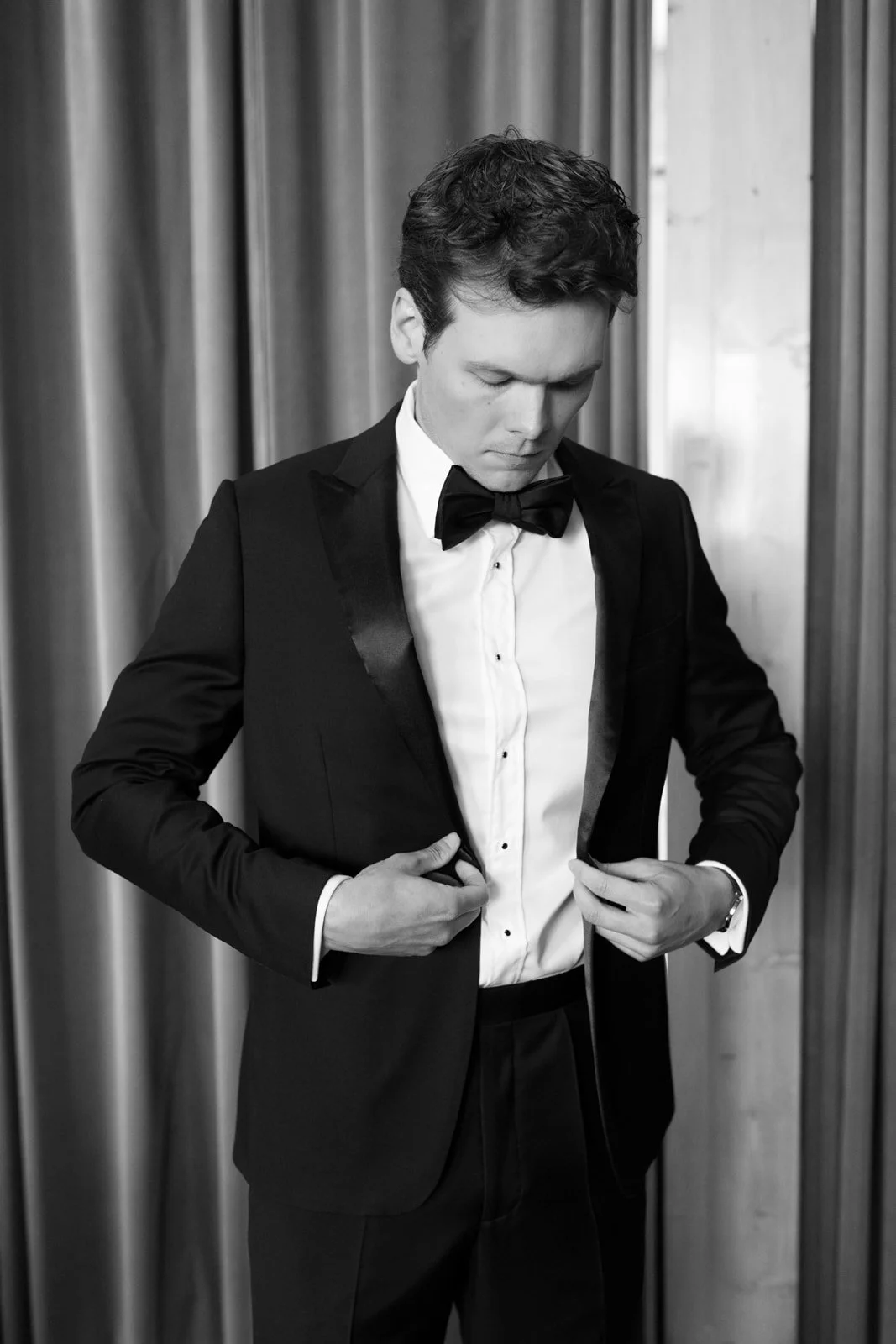 A young man in a tuxedo adjusting his suit jacket in front of curtains.